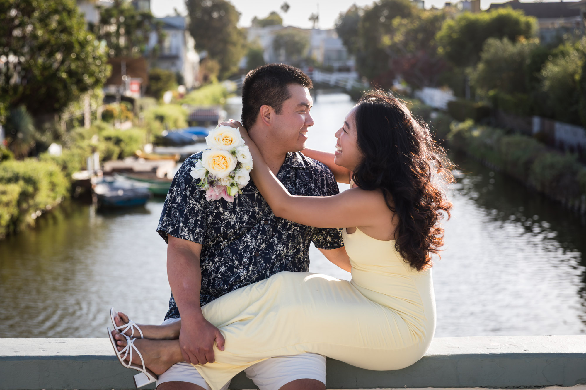 Woman wearing sleeveless yellow dress sitting in lap of man wearing blue and white patterned shirt with canals in background during a Venice Canals engagement photo shoot