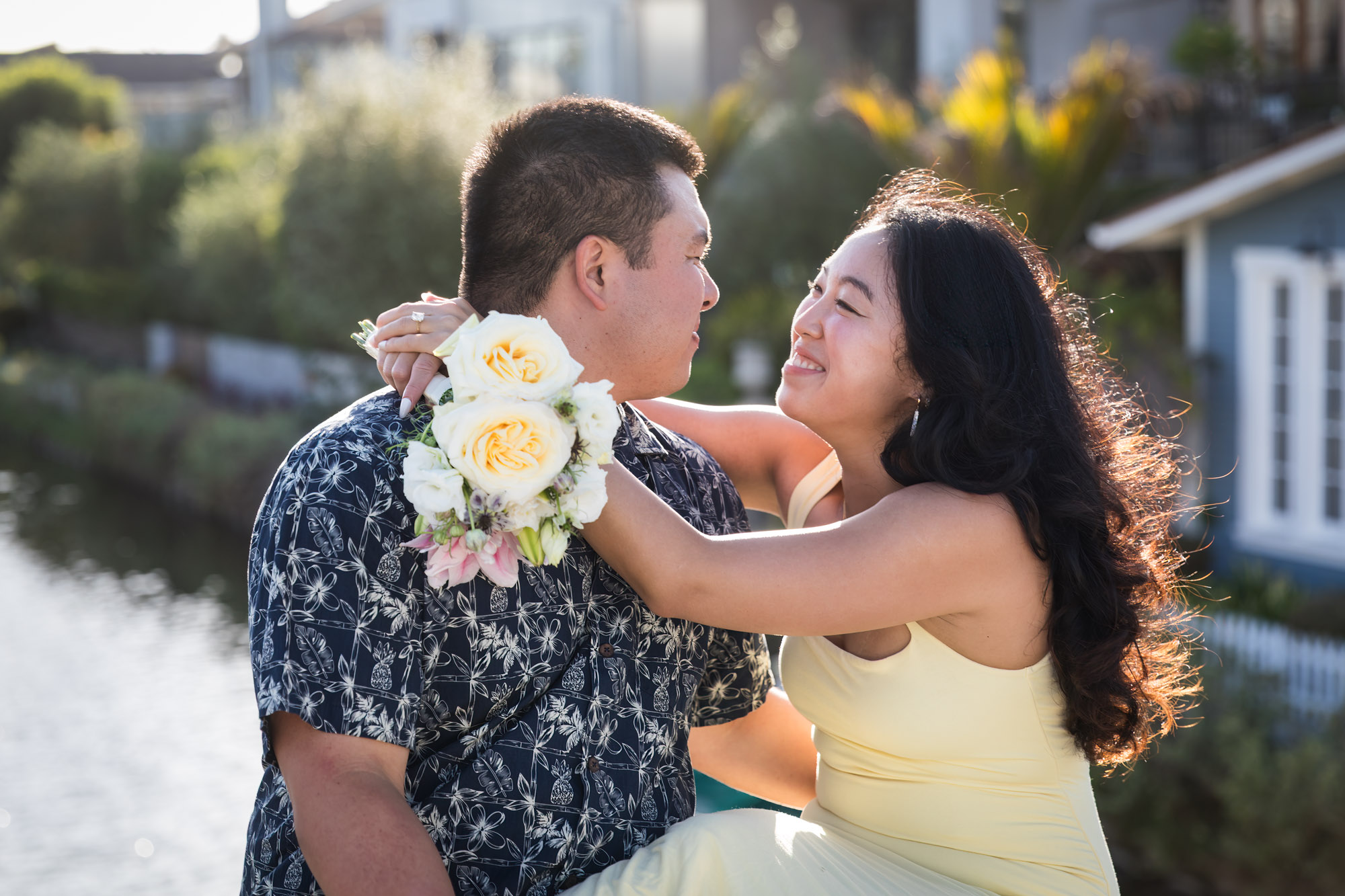 Woman wearing sleeveless yellow dress sitting in lap of man wearing blue and white patterned shirt with canals in background during a Venice Canals engagement photo shoot