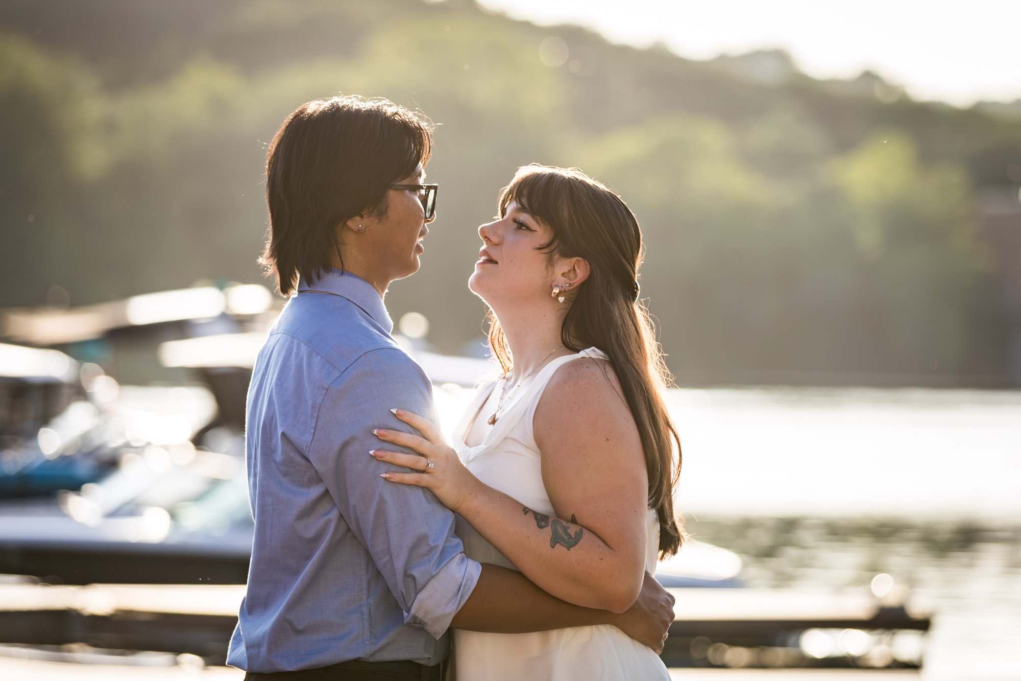 Man wearing blue shirt hugging woman wearing short, sleeveless white dress on boat dock with lake in background