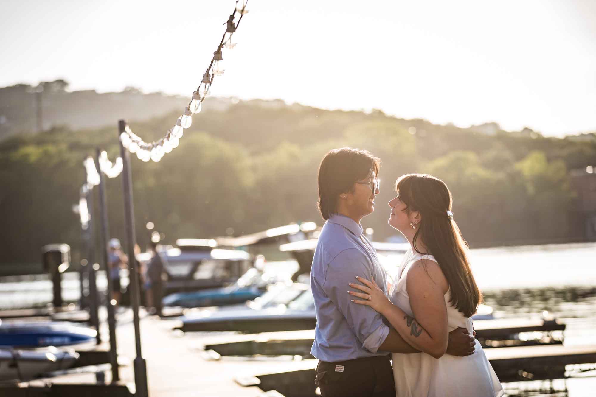 Man wearing blue shirt hugging woman wearing short, sleeveless white dress on boat dock with lake in background