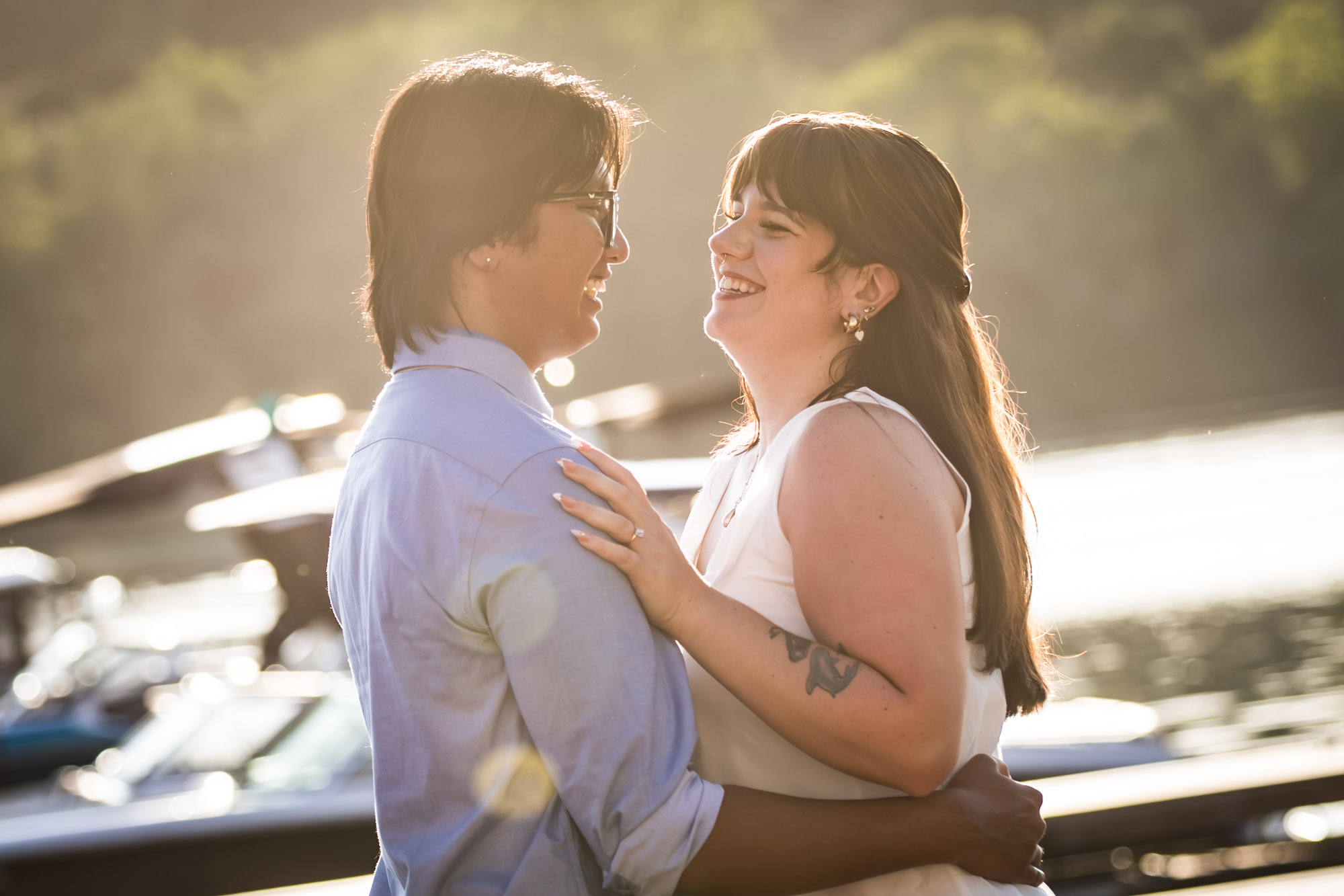 Man wearing blue shirt hugging woman wearing short, sleeveless white dress on boat dock with lake in background