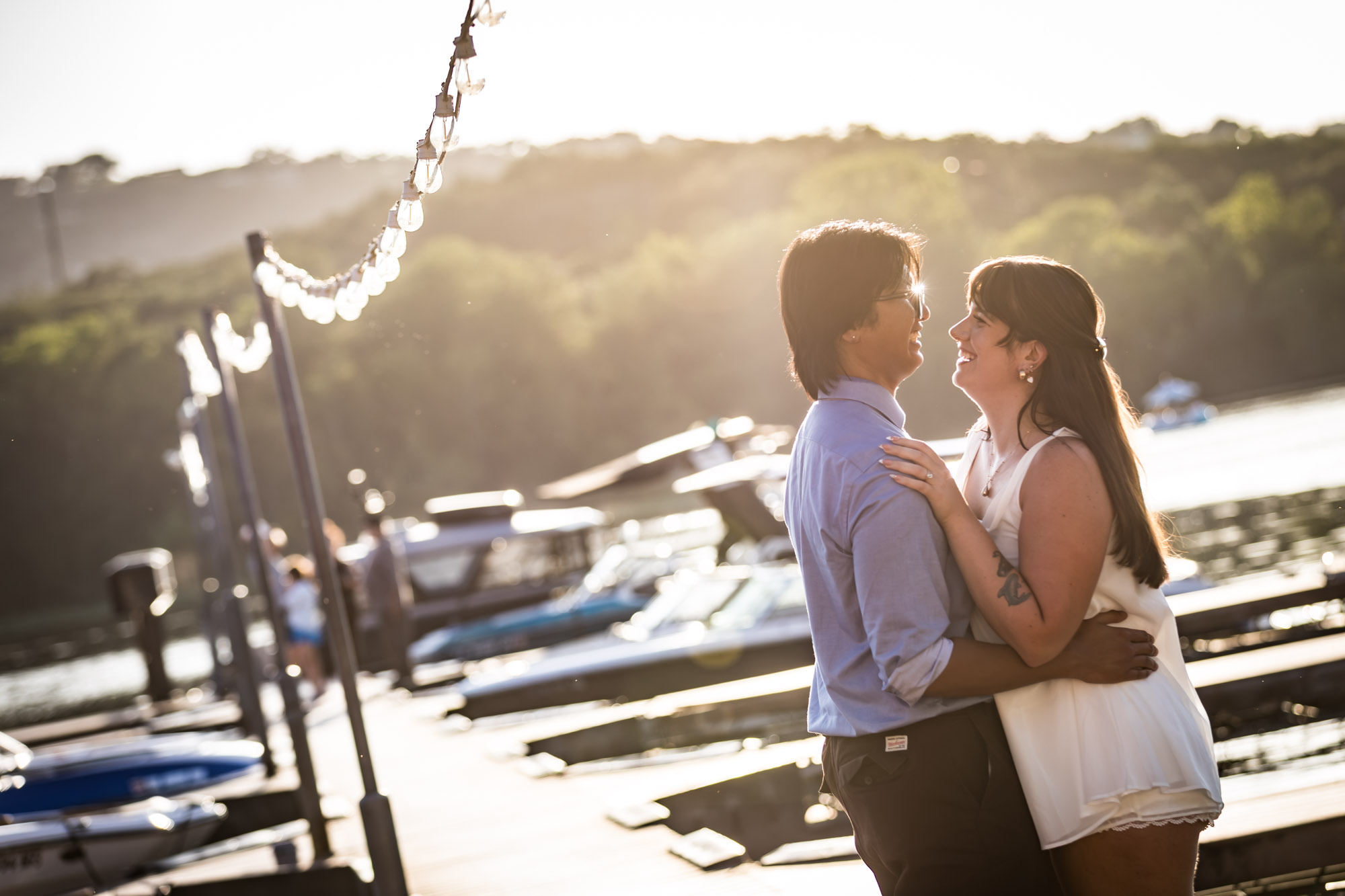 Man wearing blue shirt hugging woman wearing short, sleeveless white dress on boat dock with lake in background