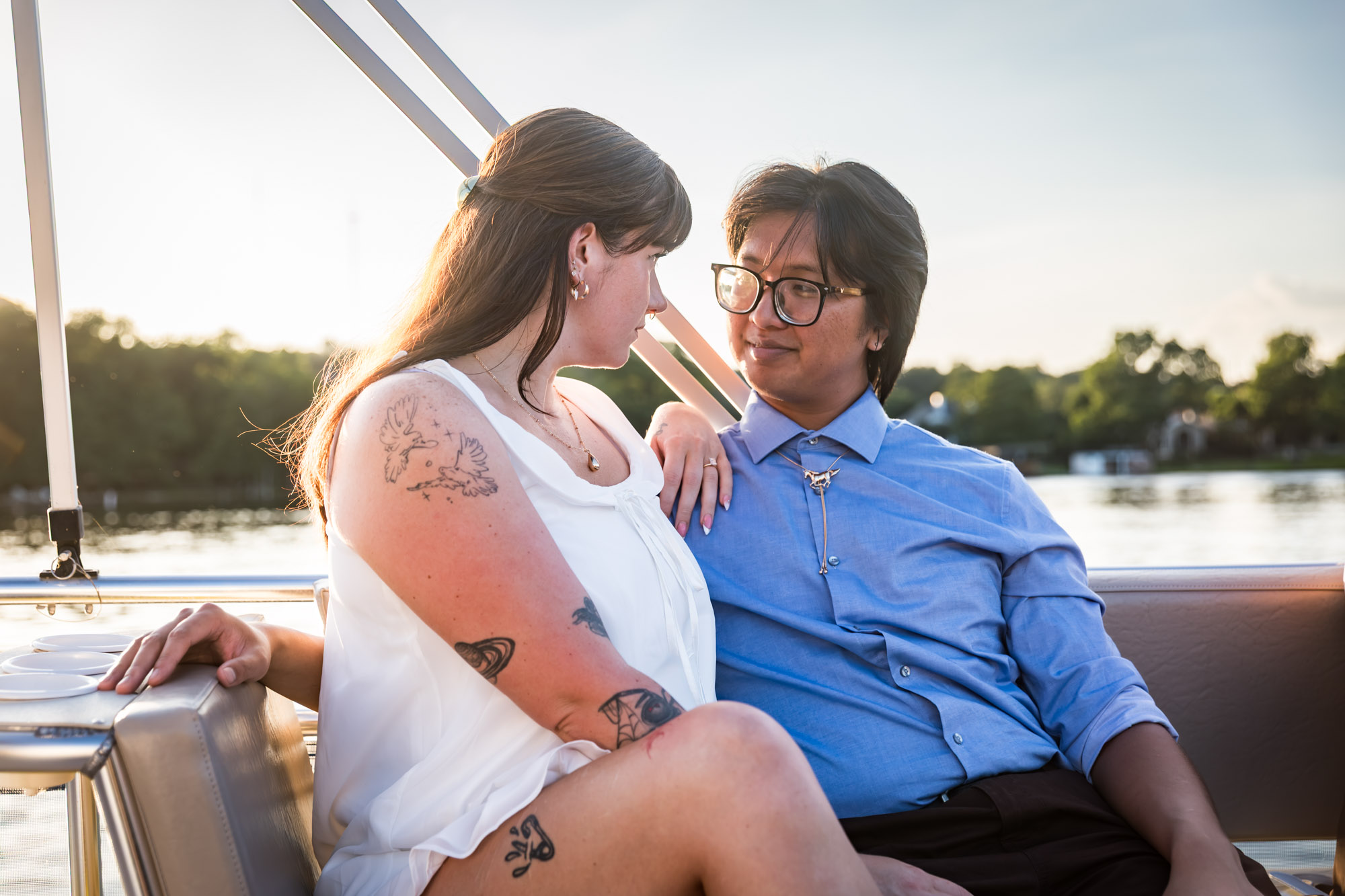 Man wearing blue shirt sitting to the right of woman wearing sleeveless, white dress on a boat with lake in the background