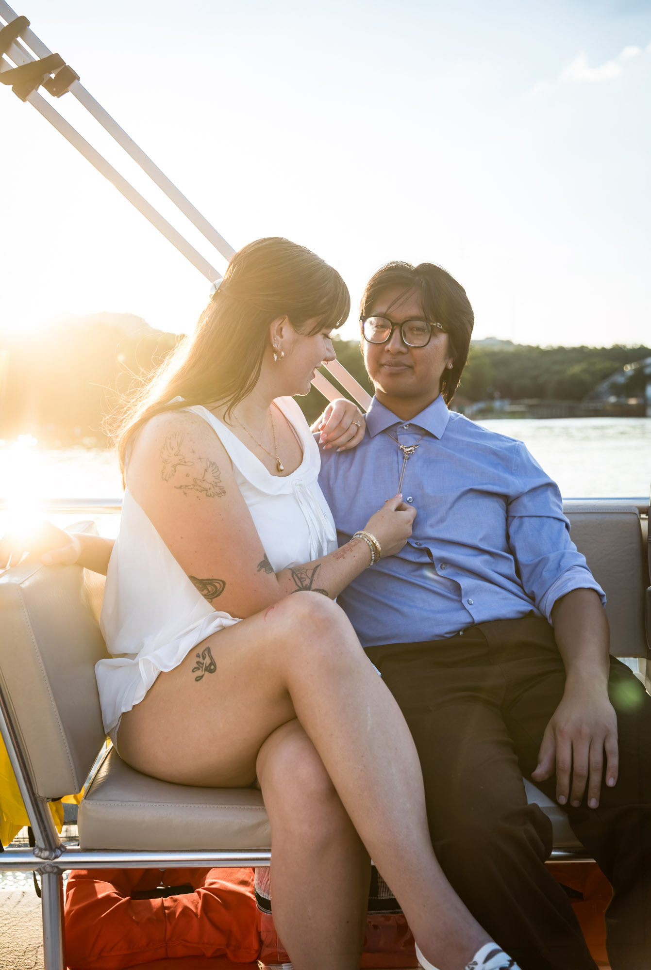 Man wearing blue shirt sitting to the right of woman wearing sleeveless, white dress on a boat with lake in the background