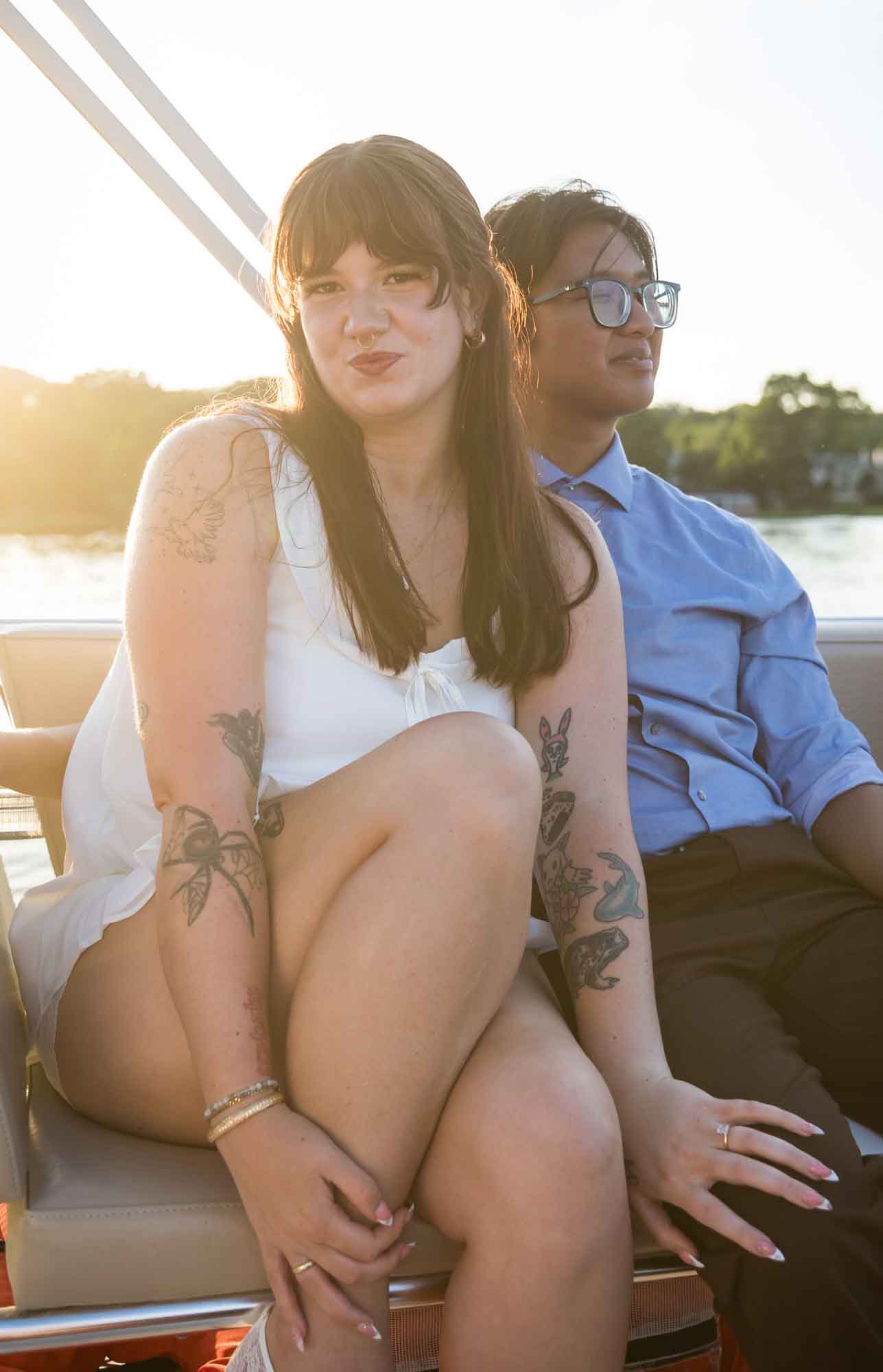 Man wearing blue shirt sitting to the right of woman wearing sleeveless, white dress on a boat with lake in the background