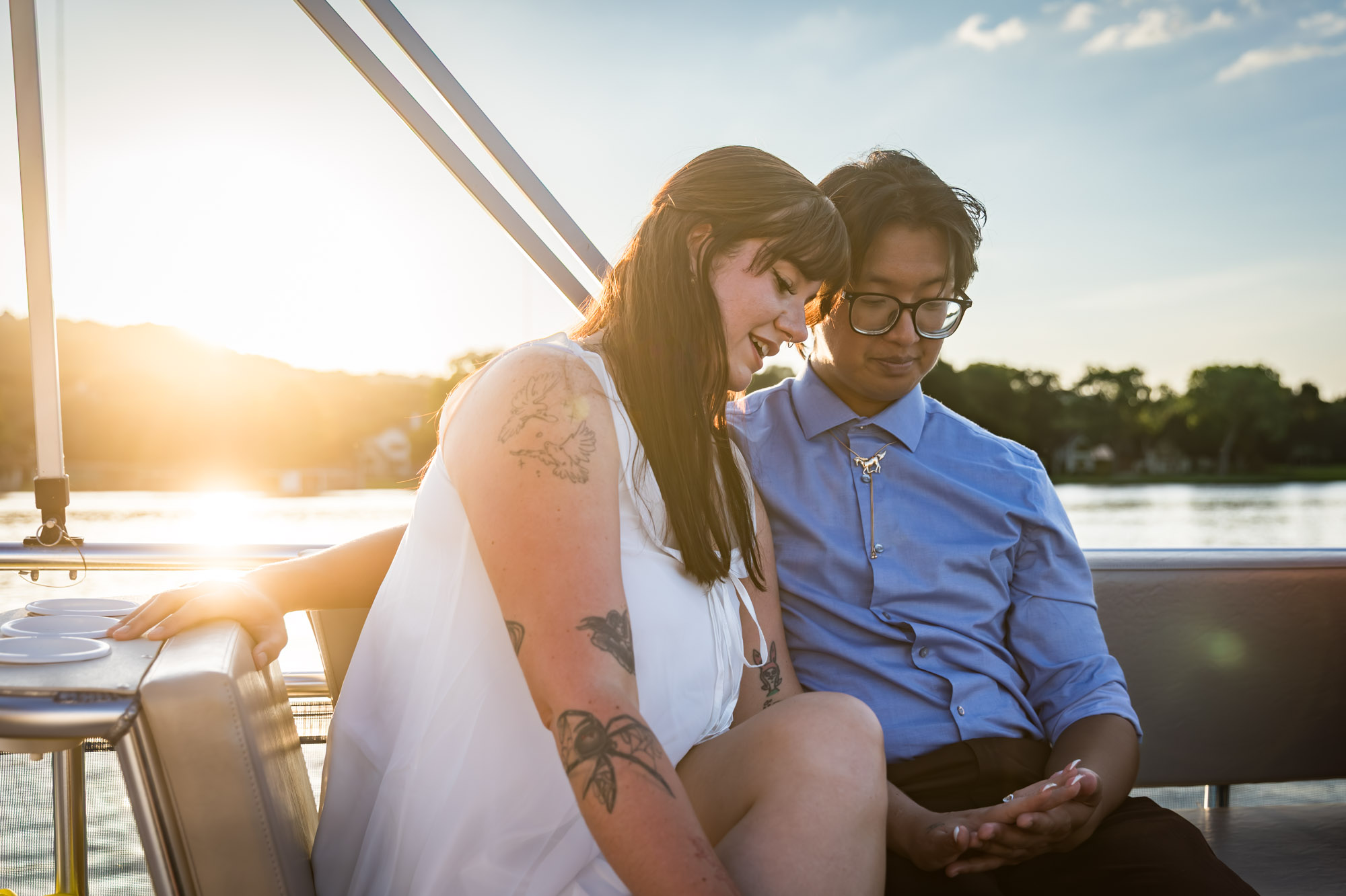 Man wearing blue shirt sitting to the right of woman wearing sleeveless, white dress on a boat with lake in the background looking at her hand showing engagement ring