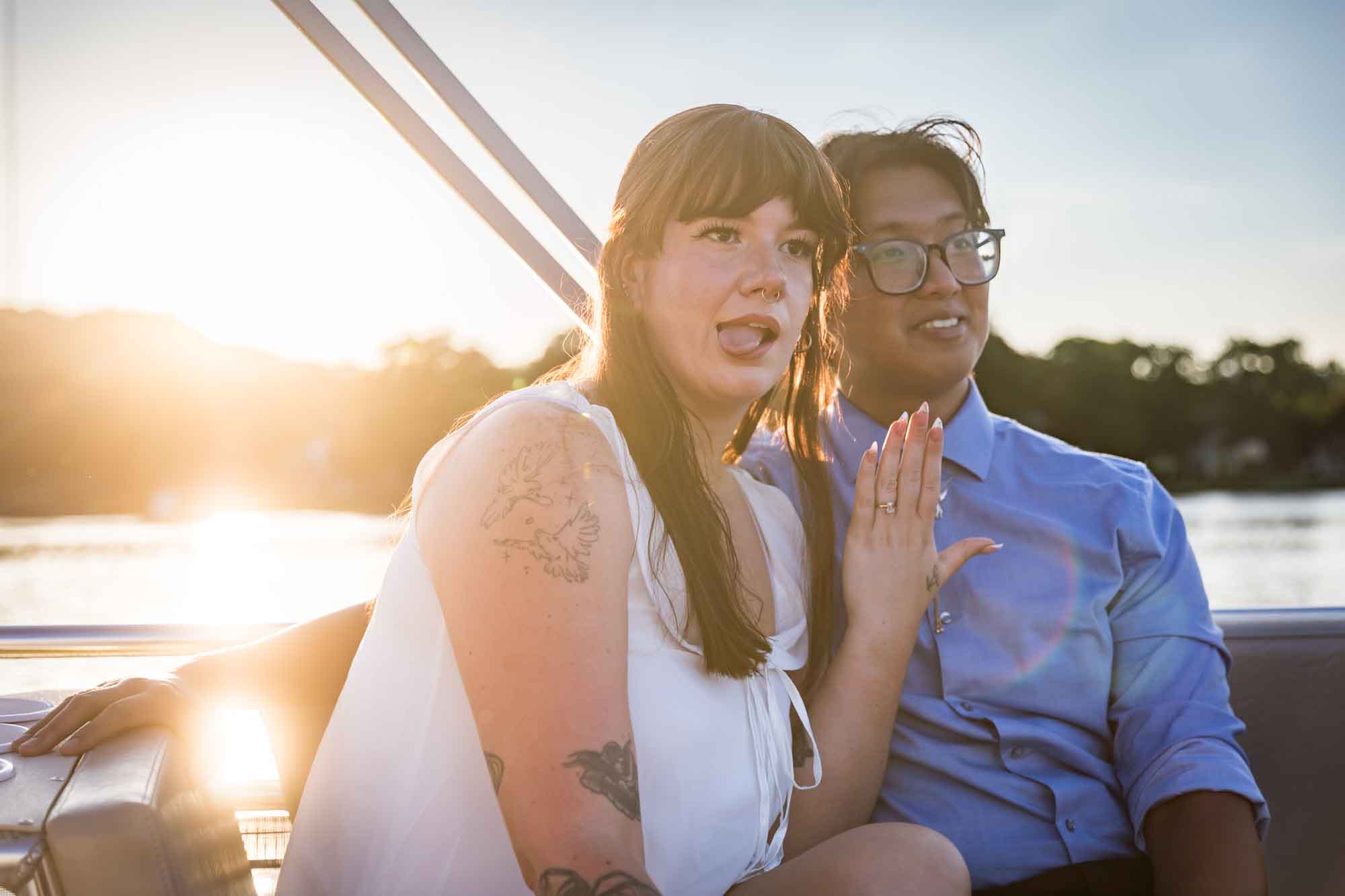 Man wearing blue shirt sitting to the right of woman wearing sleeveless, white dress showing off engagement ring on her hand