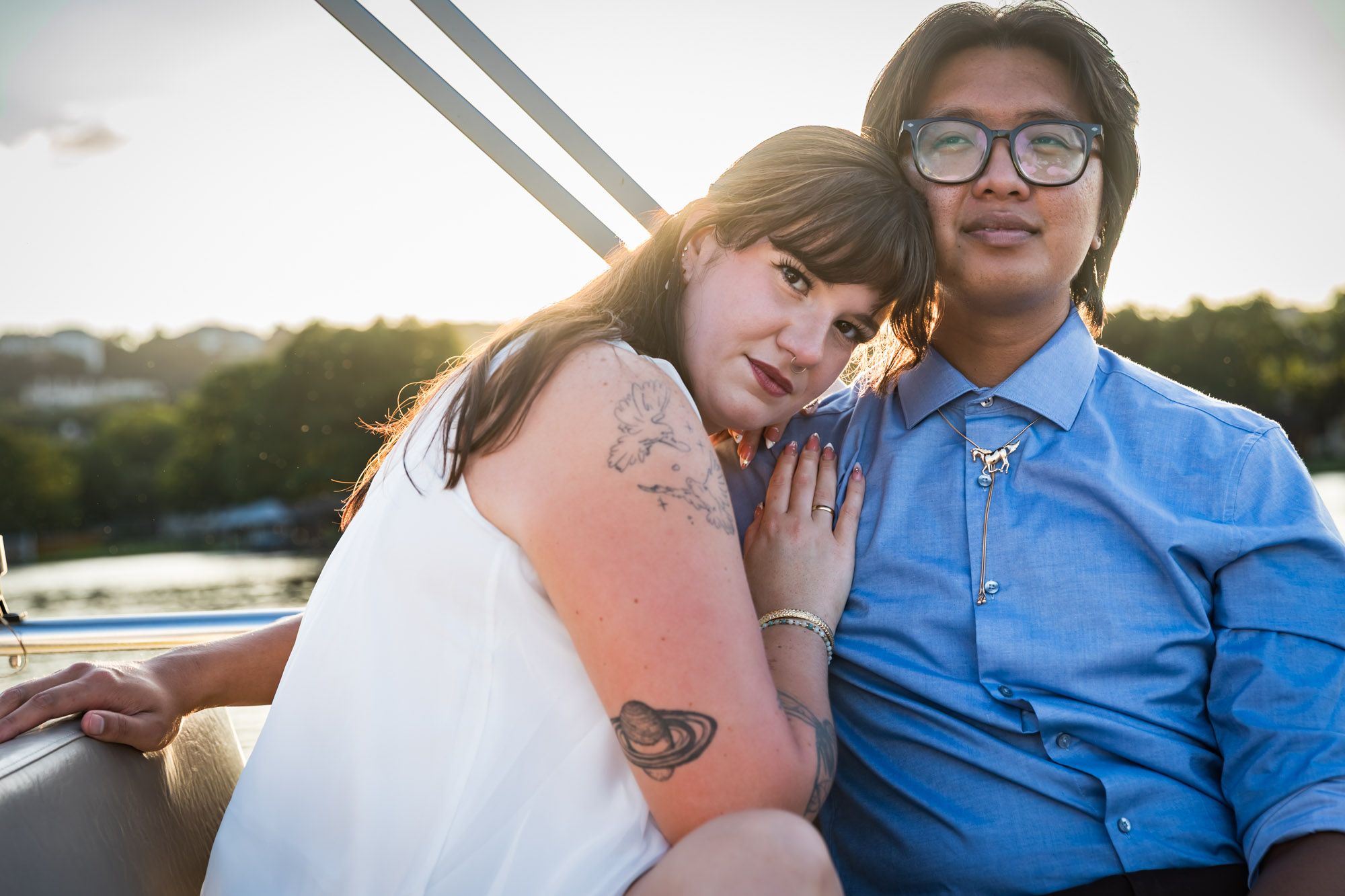 Man wearing blue shirt sitting to the right of woman wearing sleeveless, white dress on a boat with lake in the background