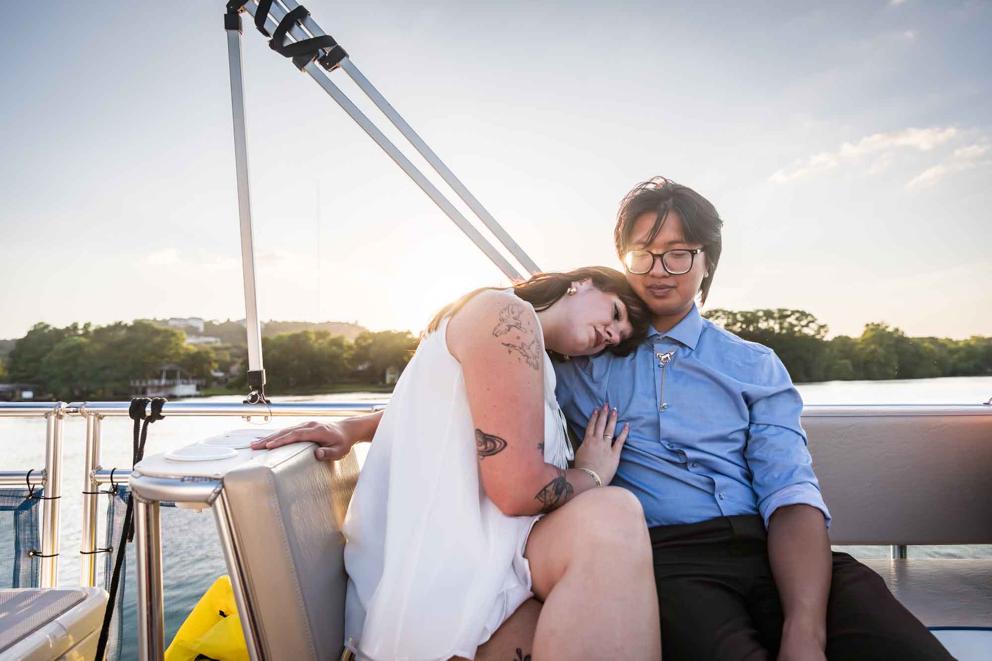 Man wearing blue shirt sitting to the right of woman wearing sleeveless, white dress on a boat with lake in the background