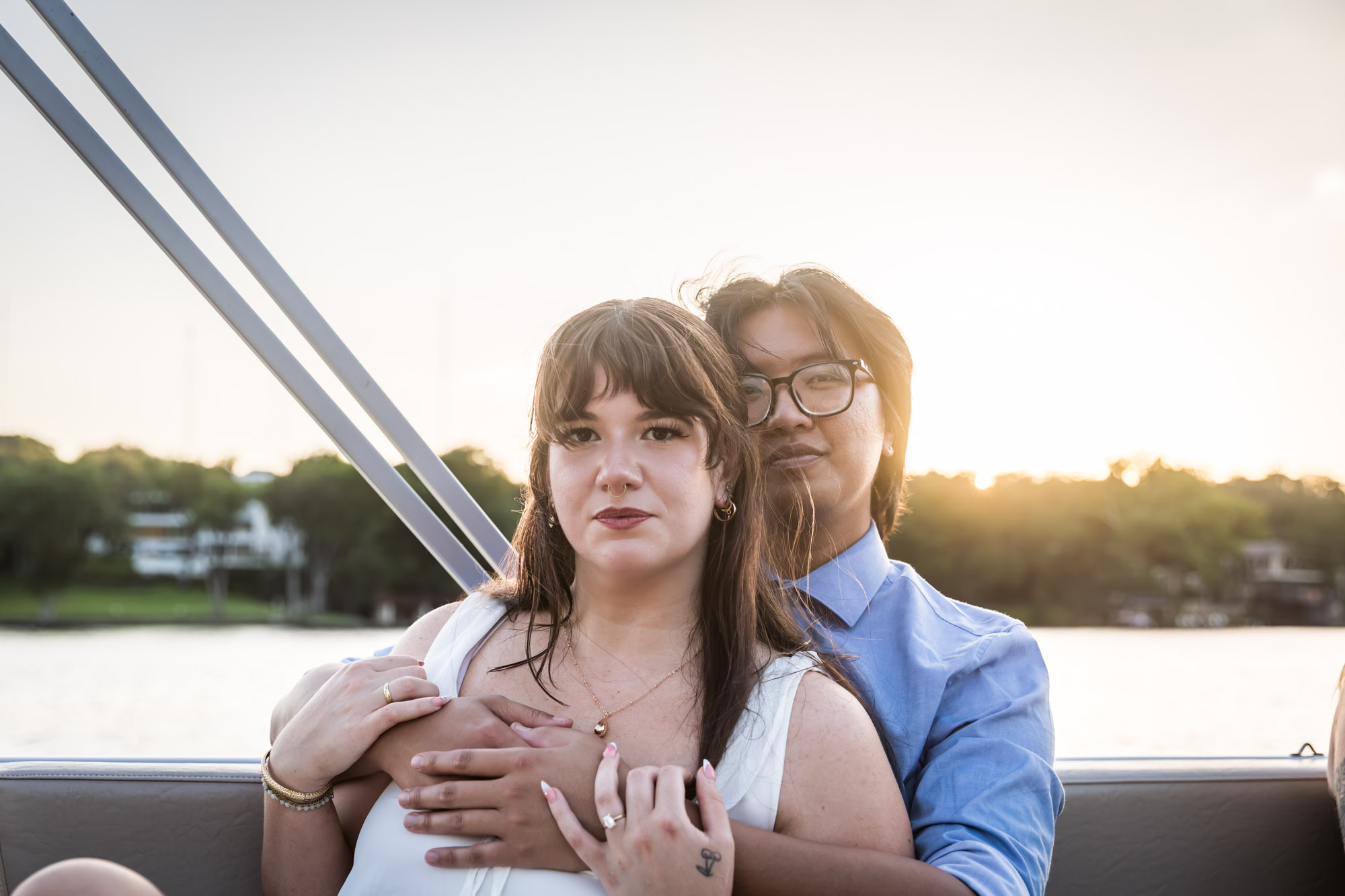 Man wearing blue shirt hugging woman wearing sleeveless, white dress on a boat with lake in the background