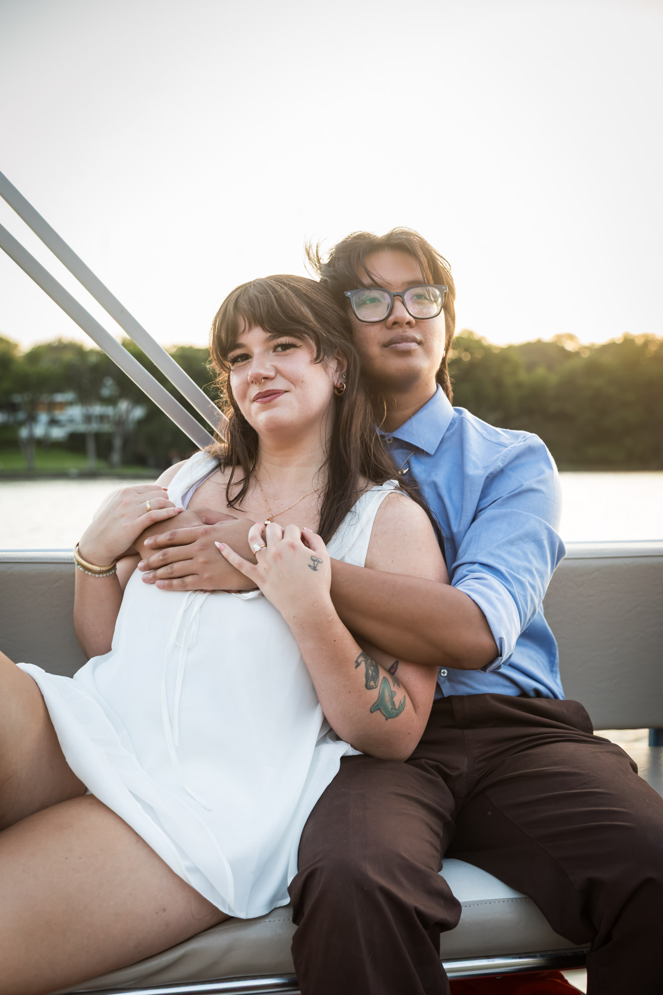Man wearing blue shirt hugging woman wearing sleeveless, white dress on a boat with lake in the background
