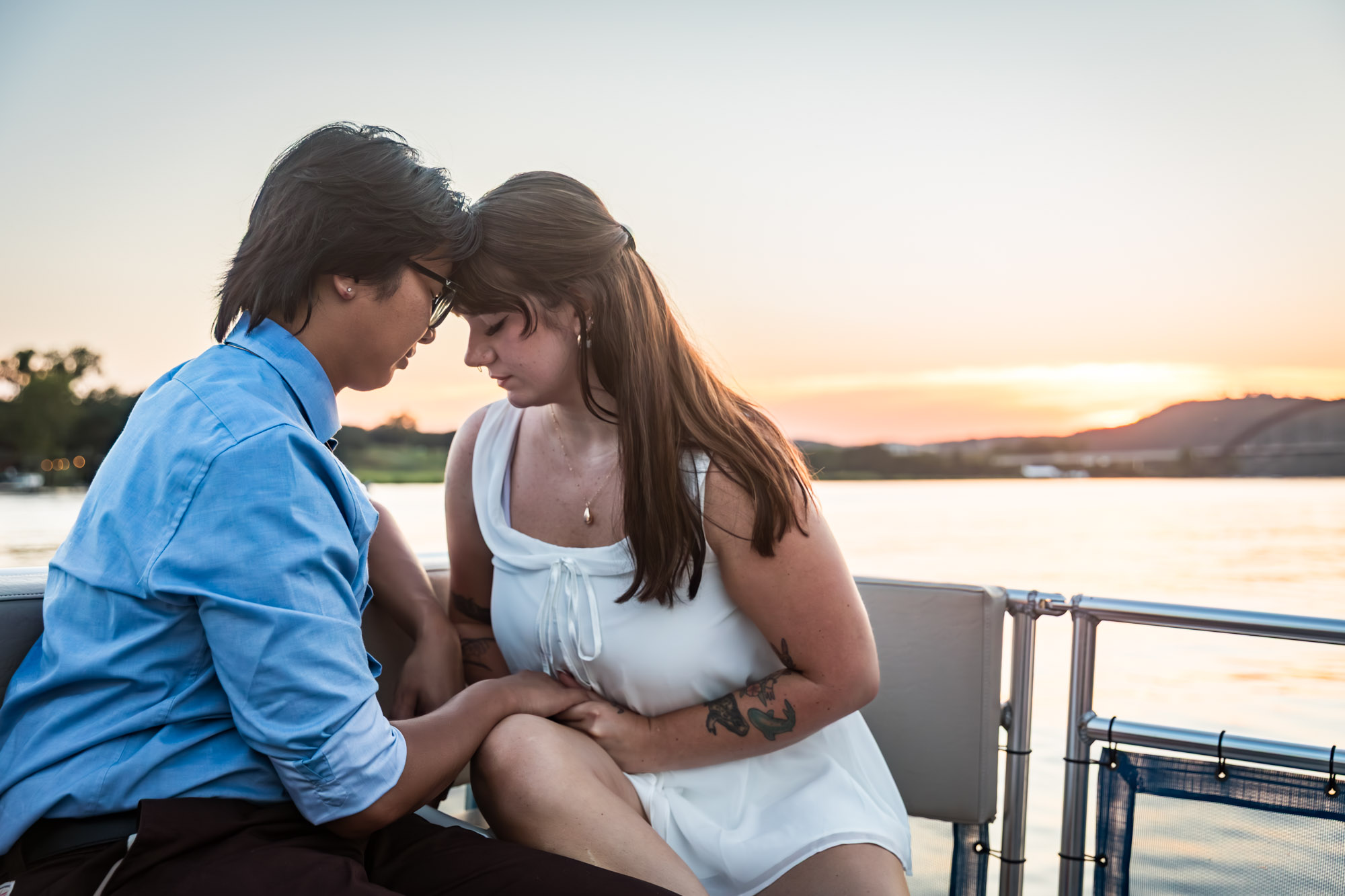 Man wearing blue shirt pressing forehead to woman wearing sleeveless, white dress on a boat with lake in the background