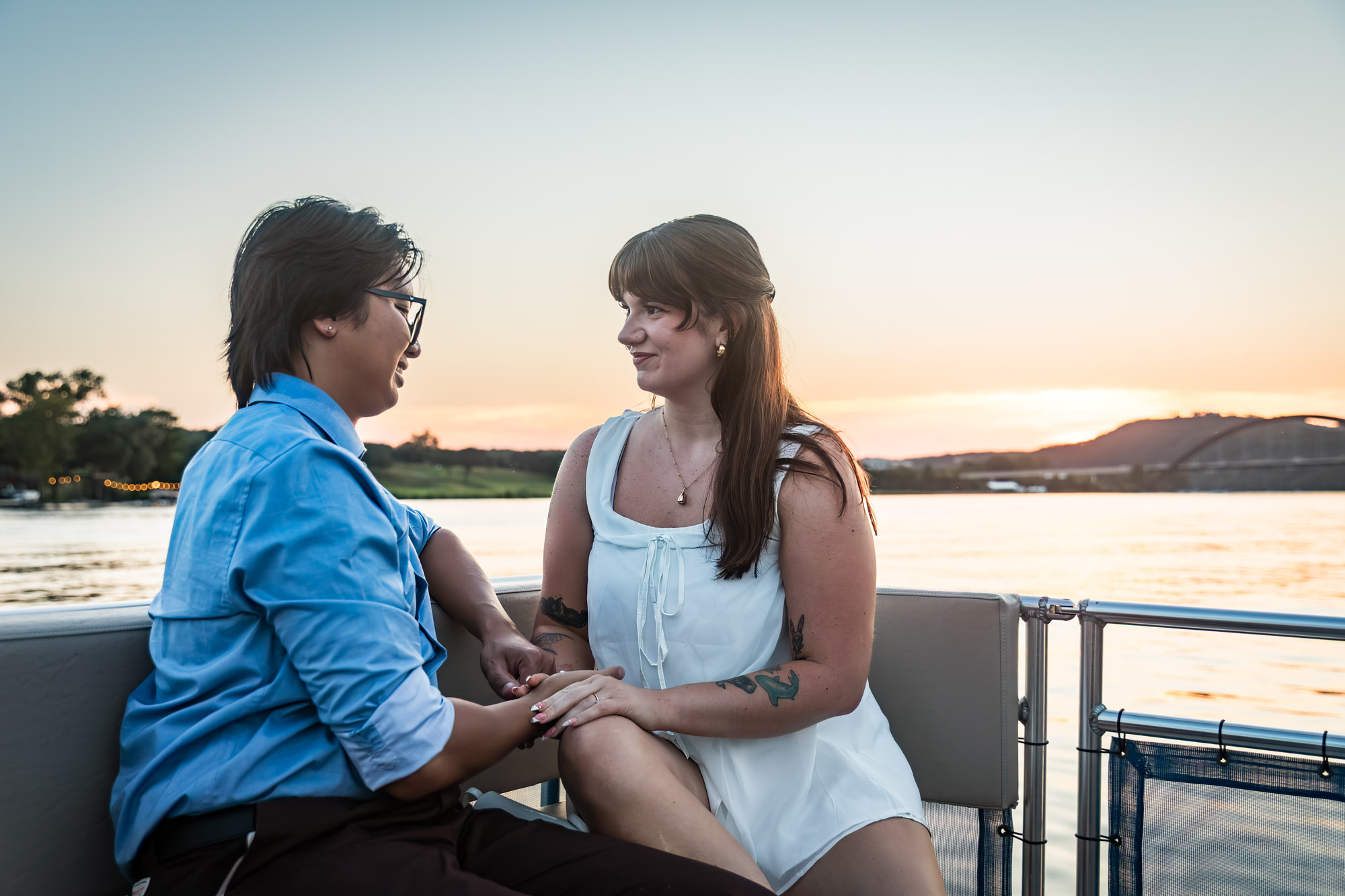Man wearing blue shirt sitting to the left of woman wearing sleeveless, white dress on a boat with lake in the background