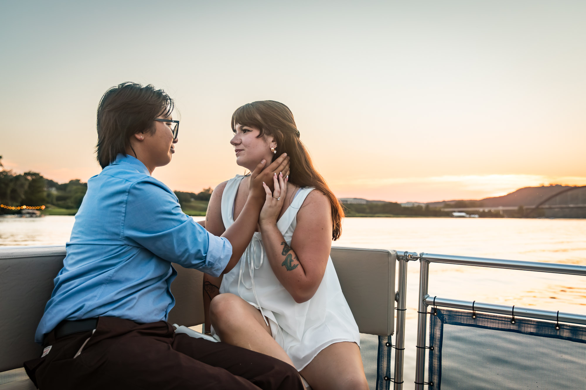 Man wearing blue shirt touching face of woman wearing sleeveless, white dress on a boat with lake in the background