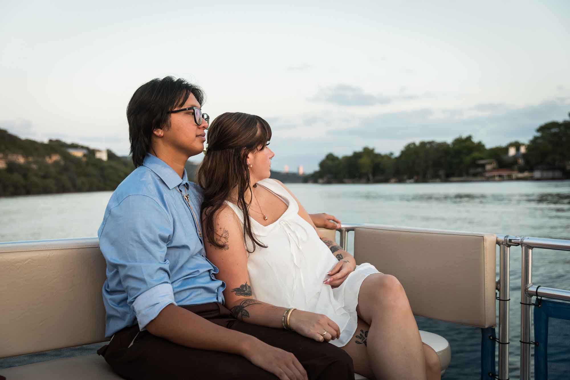 Man wearing blue shirt sitting to the left of woman wearing sleeveless, white dress on a boat with lake in the background