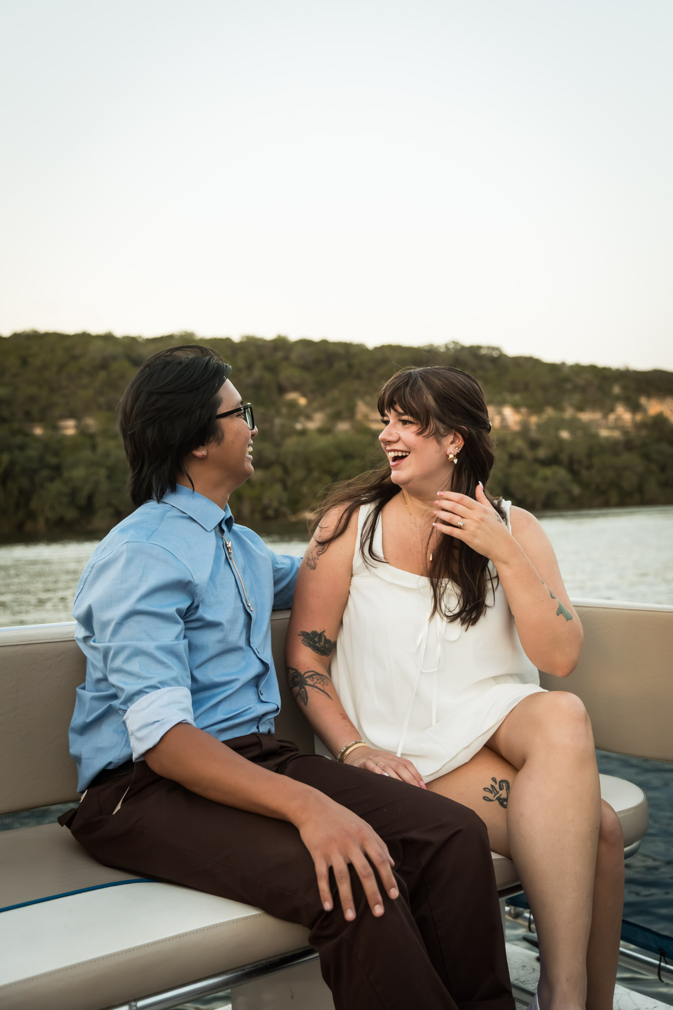 Man wearing blue shirt sitting to the left of woman wearing sleeveless, white dress on a boat with lake in the background