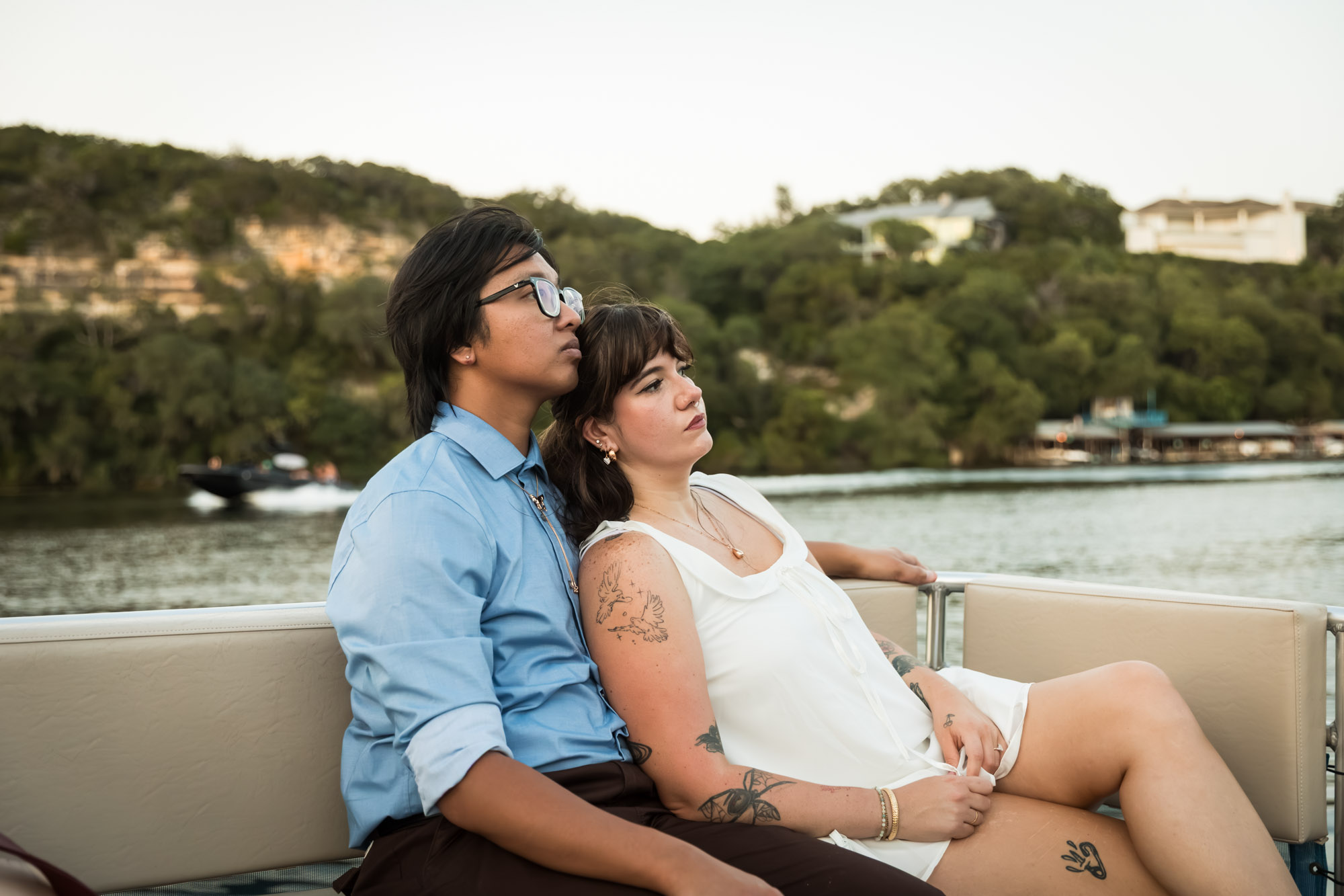 Man wearing blue shirt sitting to the left of woman wearing sleeveless, white dress on a boat with lake in the background