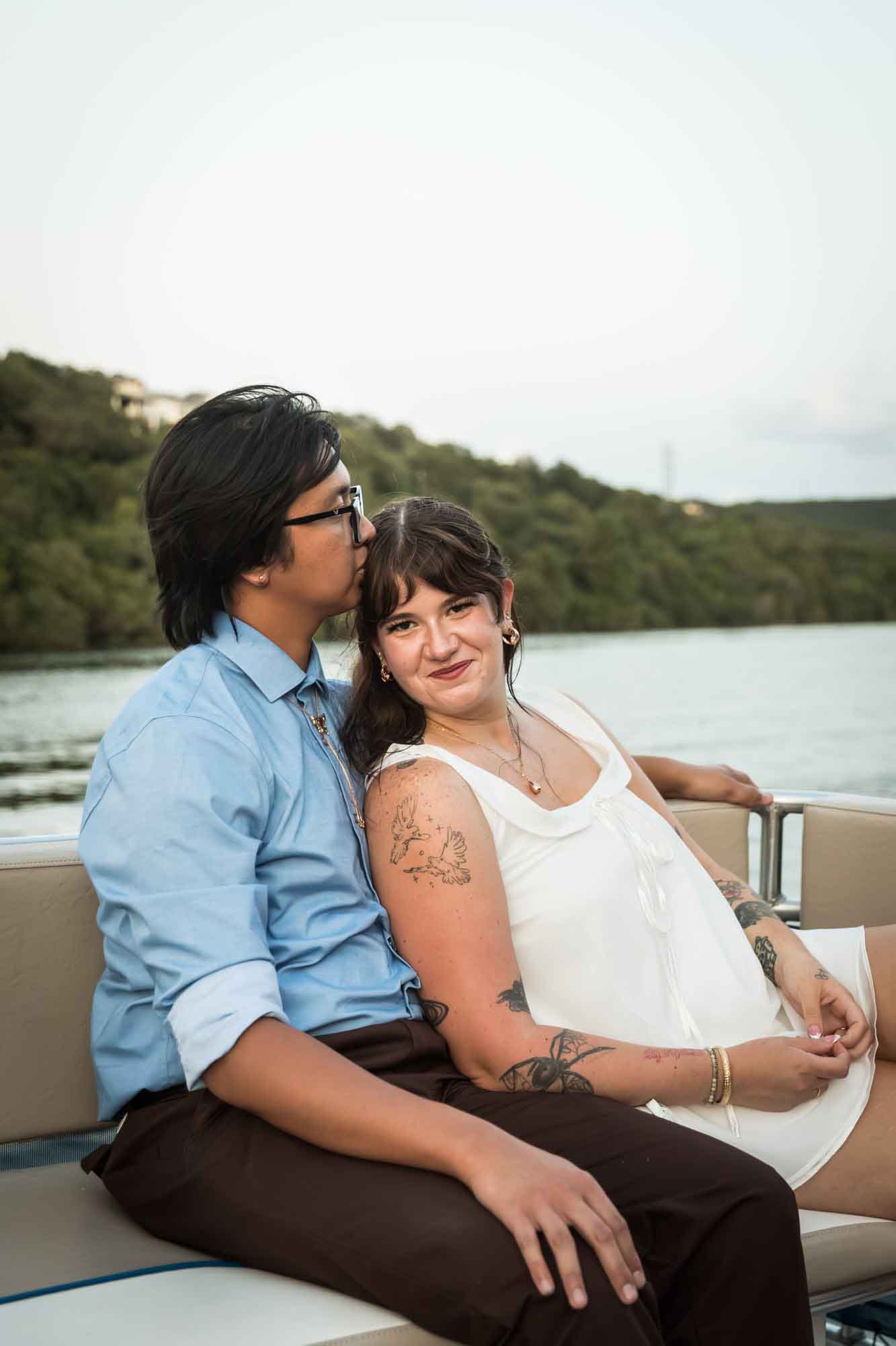 Man wearing blue shirt sitting to the left of woman wearing sleeveless, white dress on a boat with lake in the background