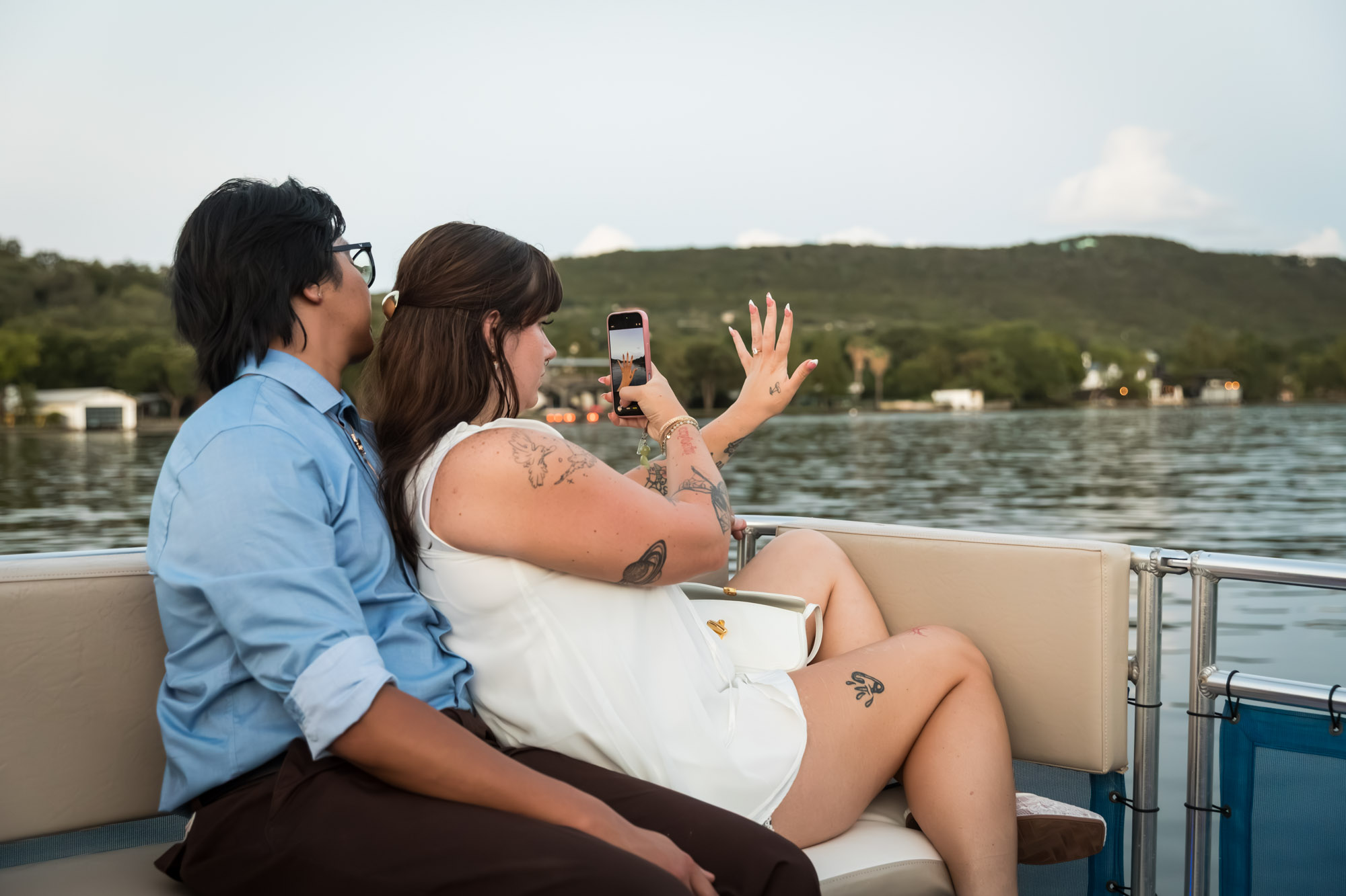 Man wearing blue shirt sitting to the left of woman wearing sleeveless, white dress taking photo of hand with phone