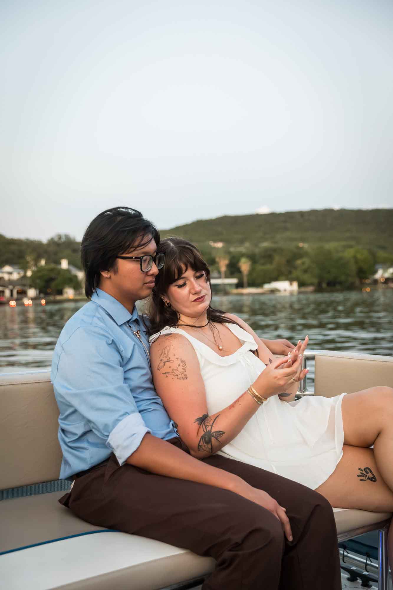 Man wearing blue shirt sitting to the left of woman wearing sleeveless, white dress on a boat with lake in the background
