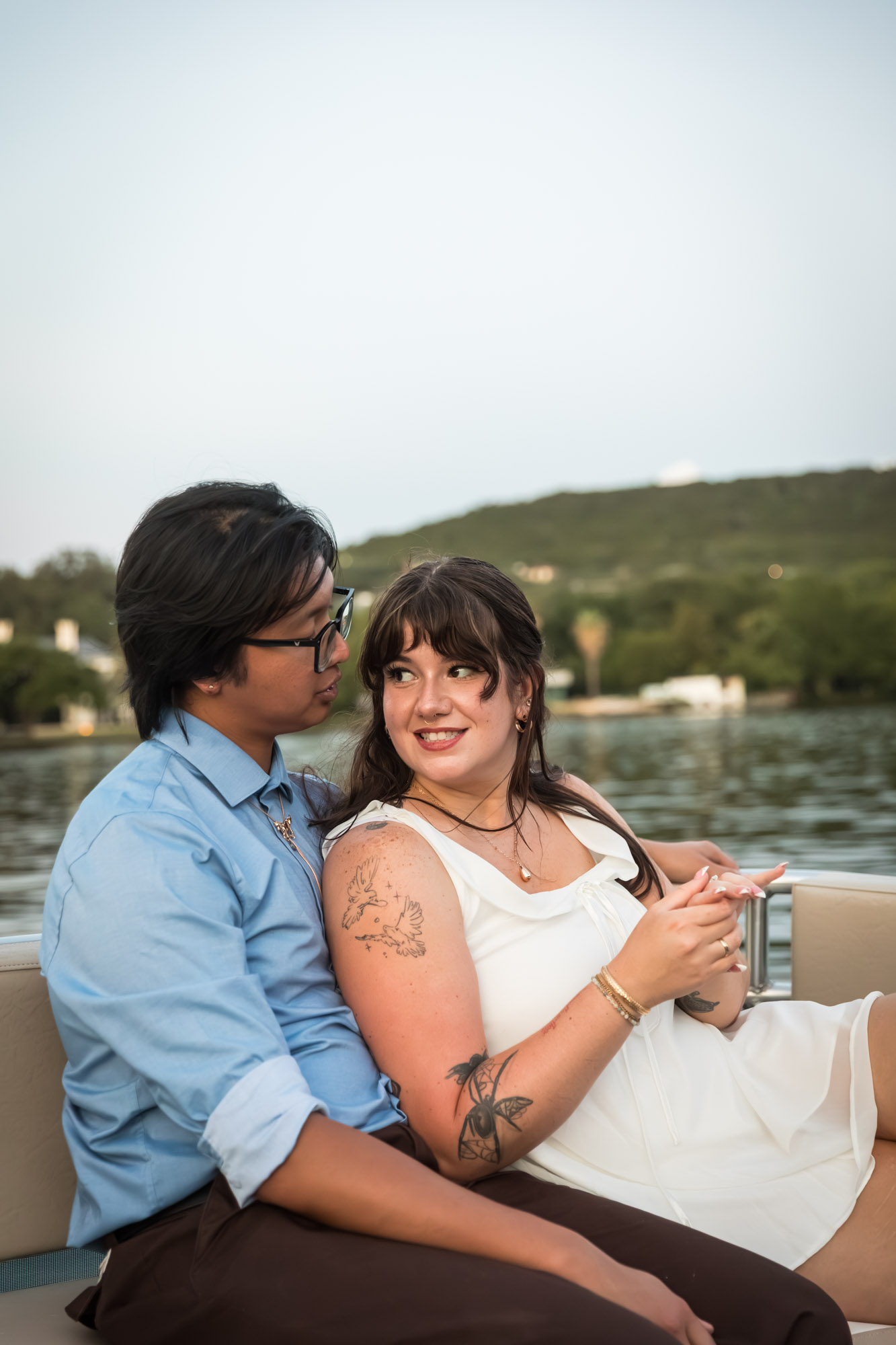 Man wearing blue shirt sitting to the left of woman wearing sleeveless, white dress on a boat with lake in the background