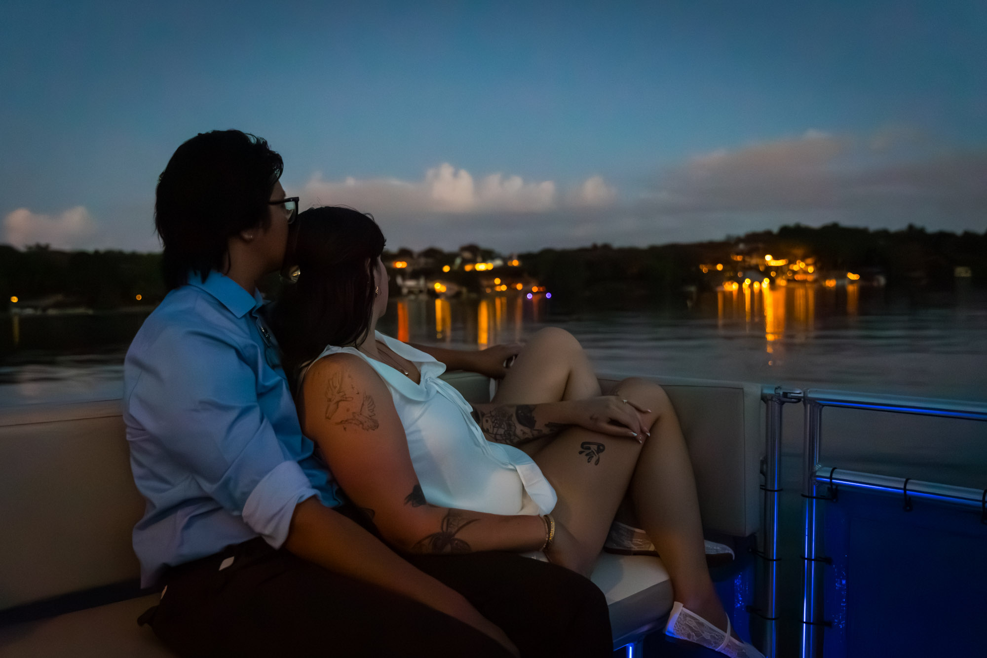 Man wearing blue shirt sitting to the left of woman wearing sleeveless, white dress on a boat with lake in the background at dusk