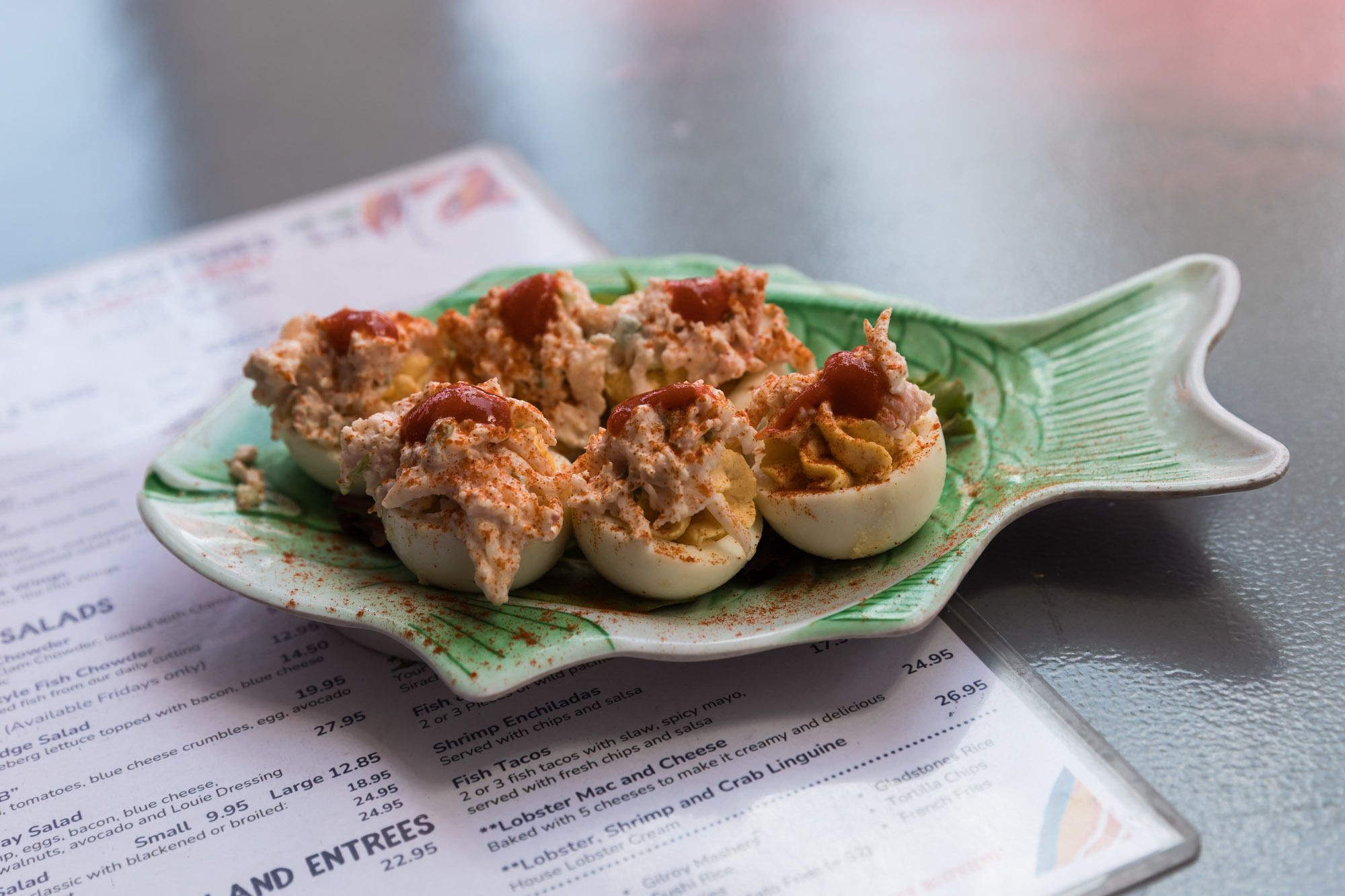 A fish-shaped, green-colored plate serving six lobster deviled eggs sitting on a menu on a table for an article on where to propose in Long Beach