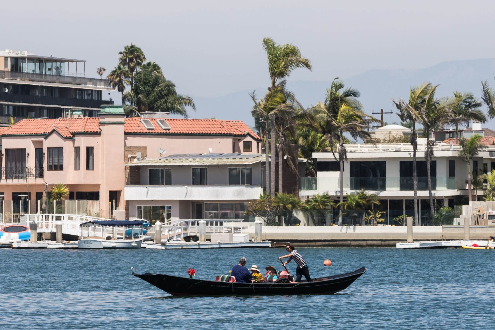 A gondolier driving a gondola with three guests on board in front of waterfront homes for an article on where to propose in Long Beach