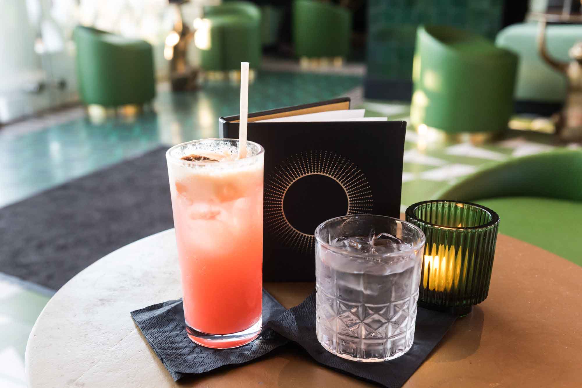 Pink mocktail in a tall glass with a short glass of water in front of a black standing menu and candle on a table for an article on where to propose in Long Beach