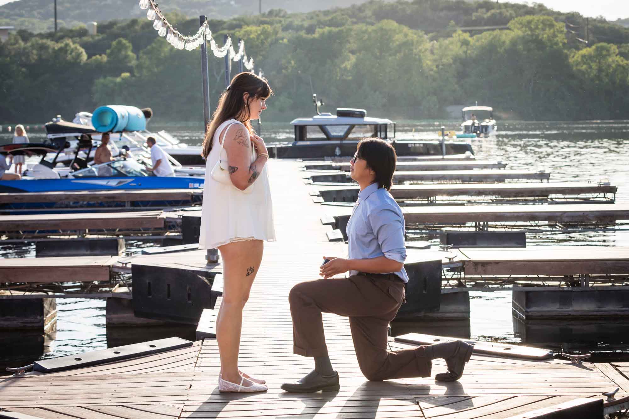 Man wearing blue shirt bending down on one knee proposing on boat dock to woman wearing short white dress