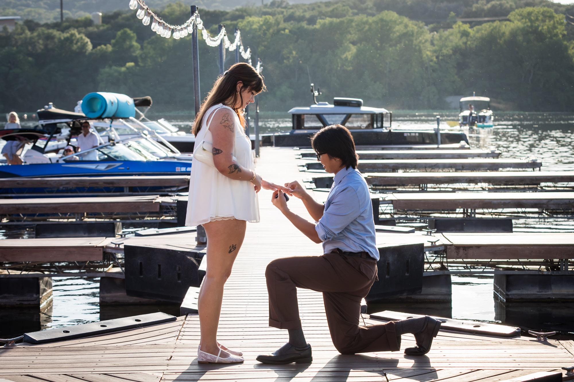 Man wearing blue shirt bending down on one knee proposing on boat dock to woman wearing short white dress