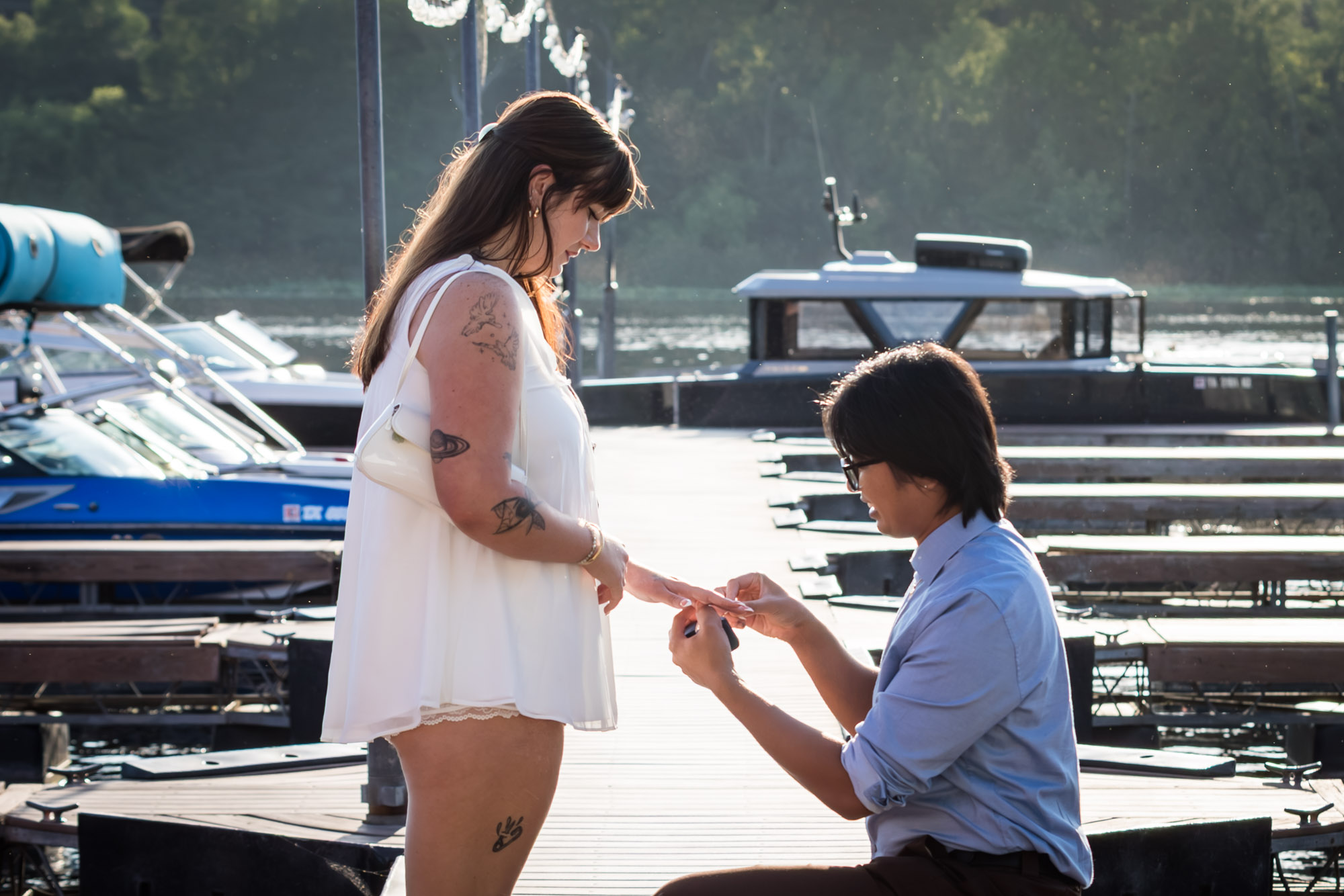 Man wearing blue shirt bending down on one knee proposing on boat dock to woman wearing short white dress