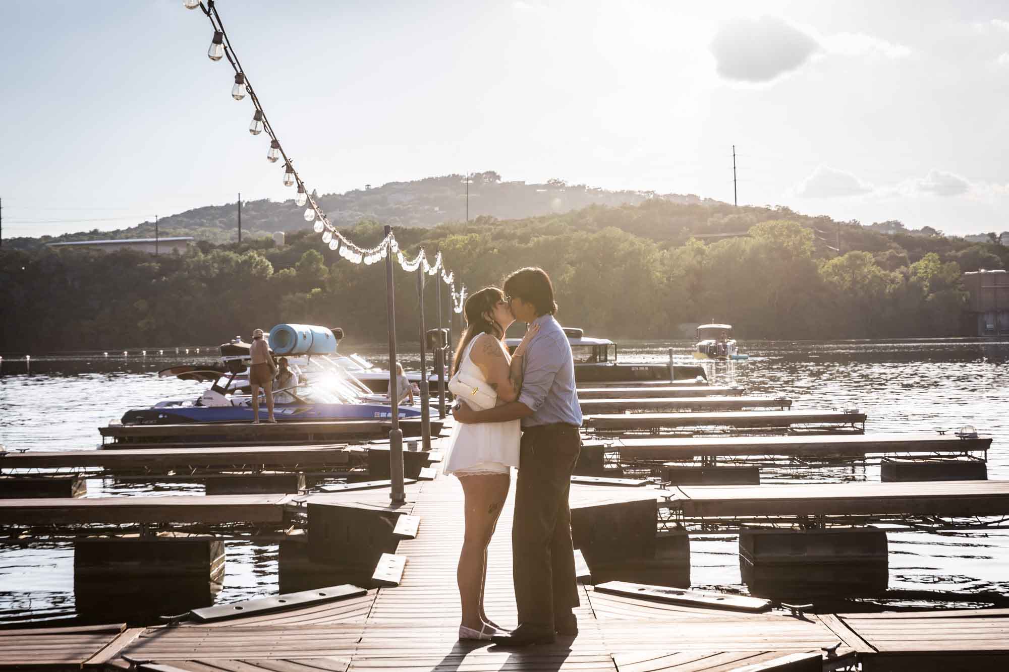Man wearing blue shirt hugging woman wearing short white dress on a boat dock in a lake with string lights above