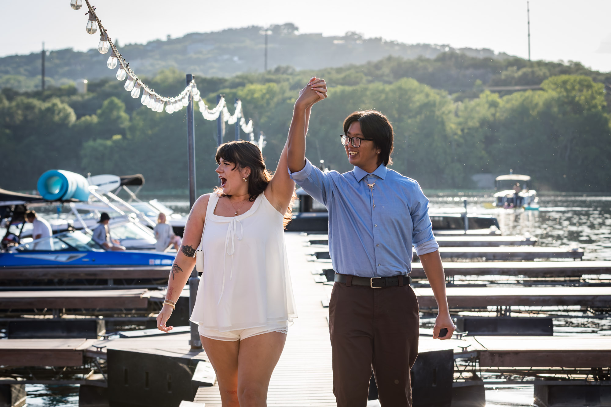 Man wearing blue shirt holding hands in the air with woman wearing short white dress on a boat dock in a lake with string lights above