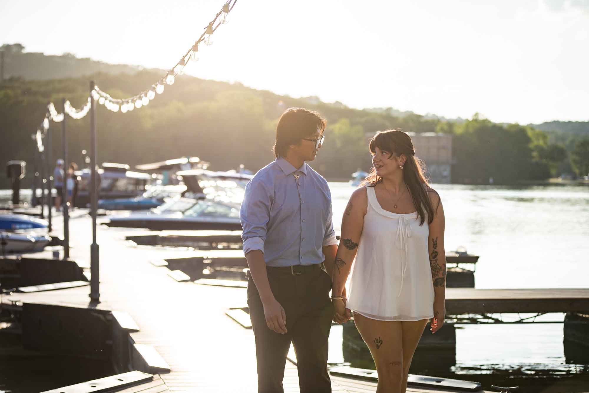 Man wearing blue shirt holding hands and walking on boat dock with woman wearing short, sleeveless white dress with lake in background