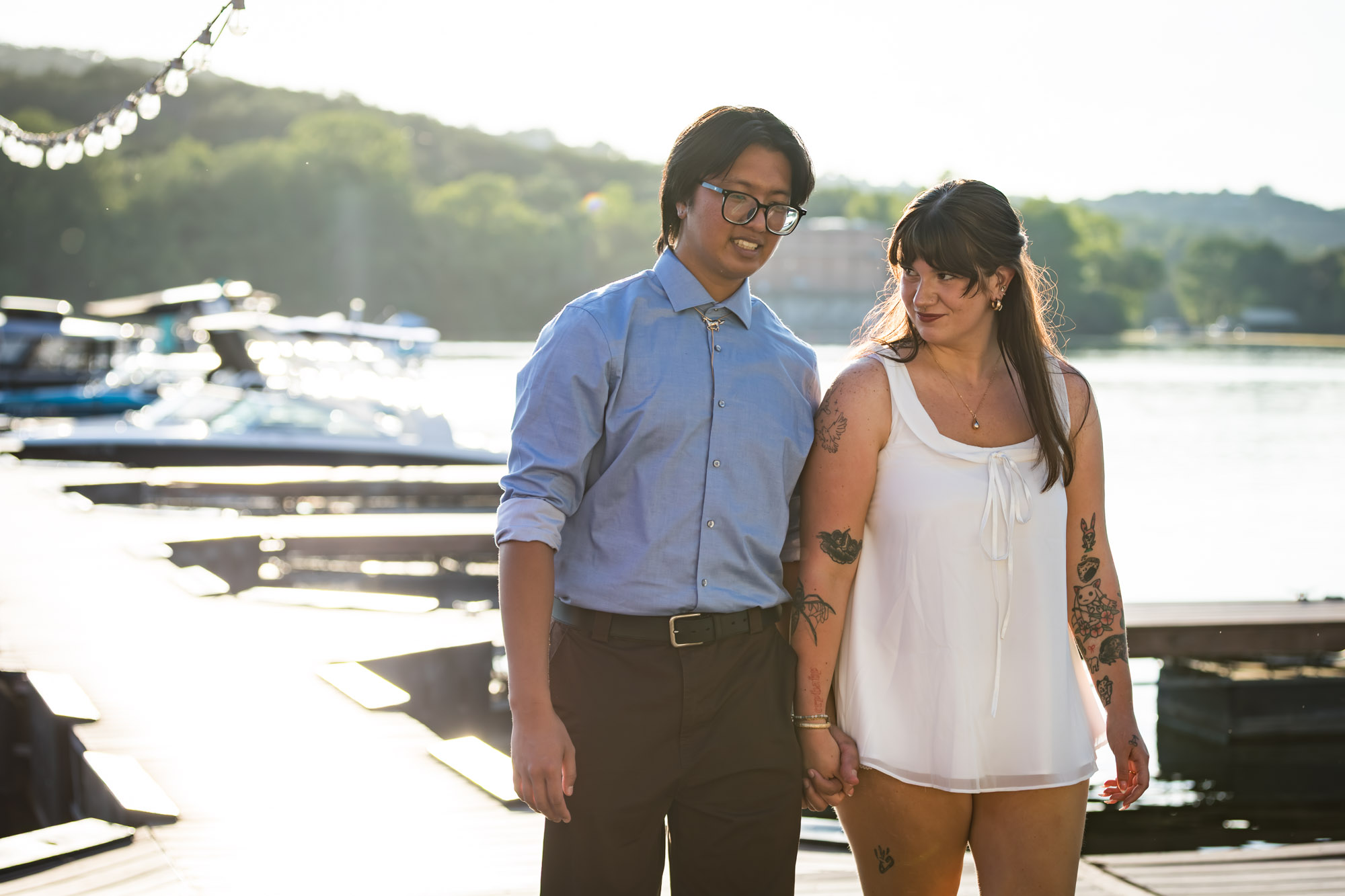 Man wearing blue shirt holding hands and walking on boat dock with woman wearing short, sleeveless white dress with lake in background