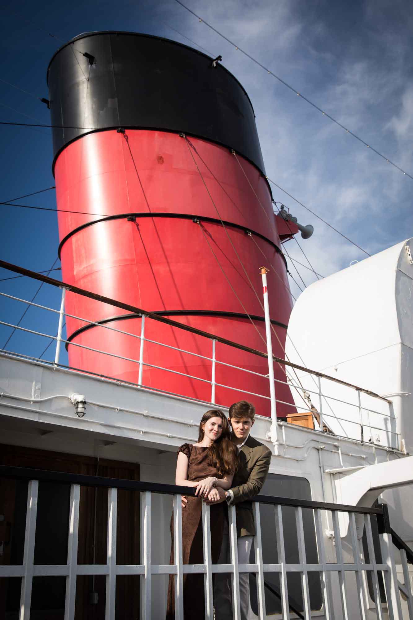Couple leaning on a white railing beneath a red and black Queen Mary funnel, a bold backdrop for a Queen Mary photo shoot