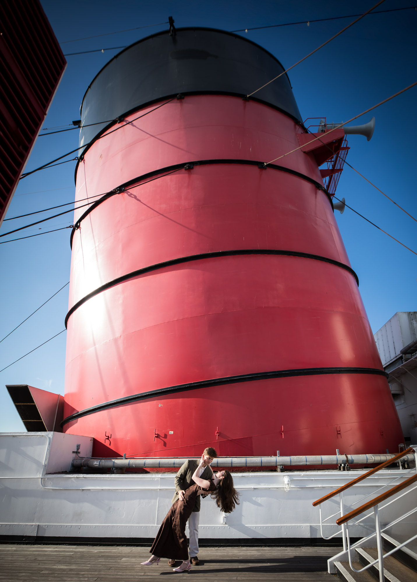 Man dipping the woman in front of a towering red funnel on the Queen Mary deck during their Queen Mary photo shoot
