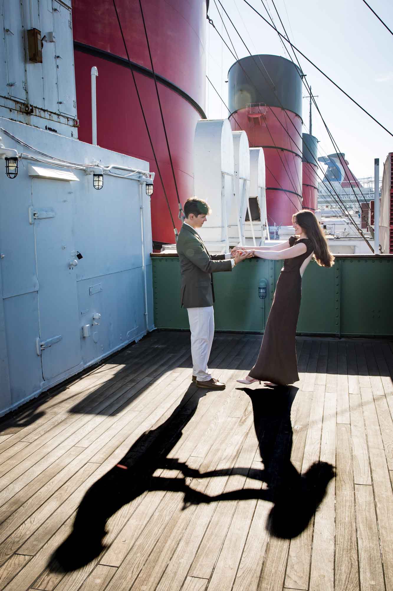Couple holding hands on the teak deck with long shadows beneath red funnels, captured during a Queen Mary photo shoot