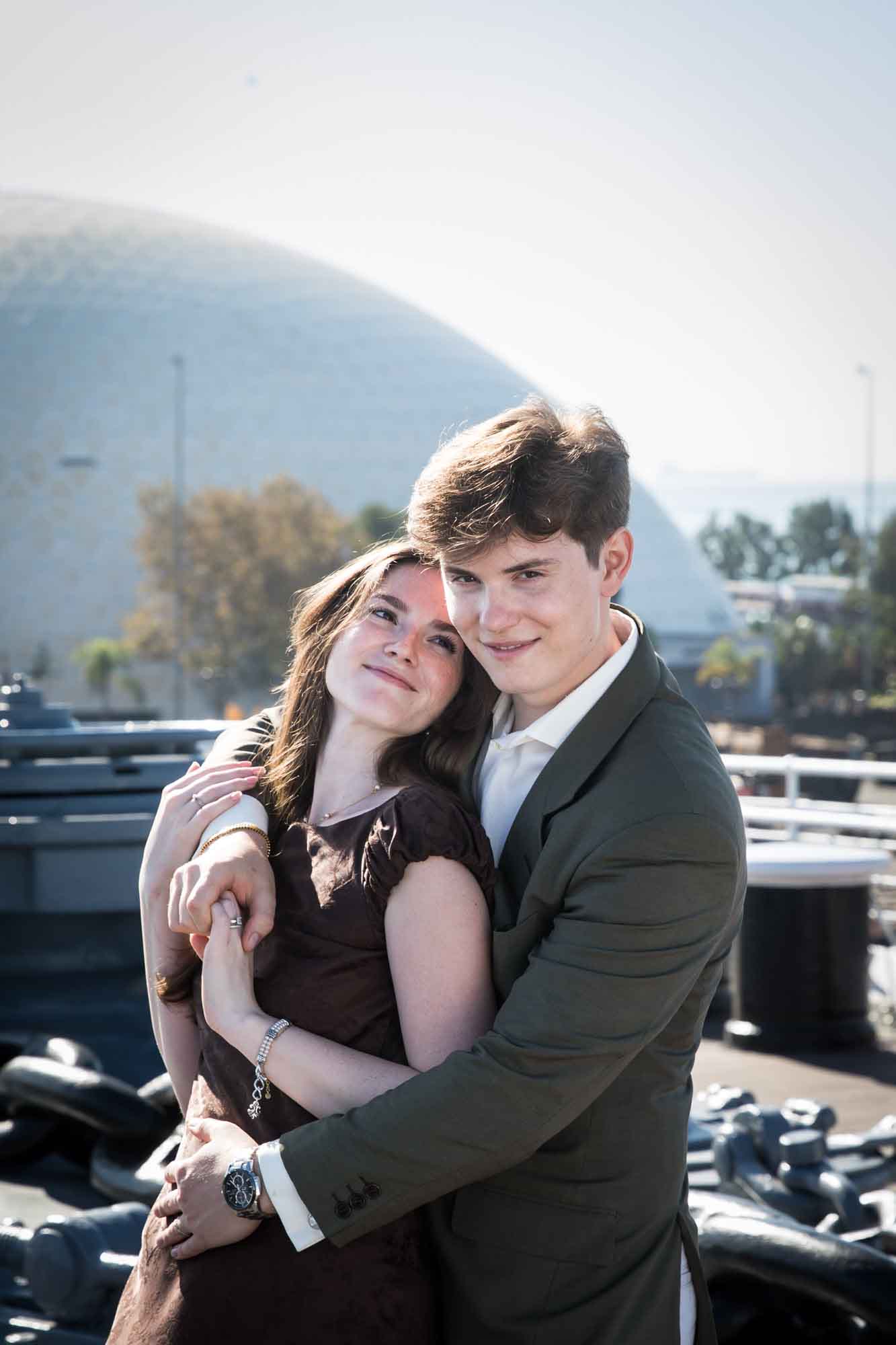 Smiling couple hugging with her head on his shoulder and the Long Beach dome behind them, Queen Mary photo shoot