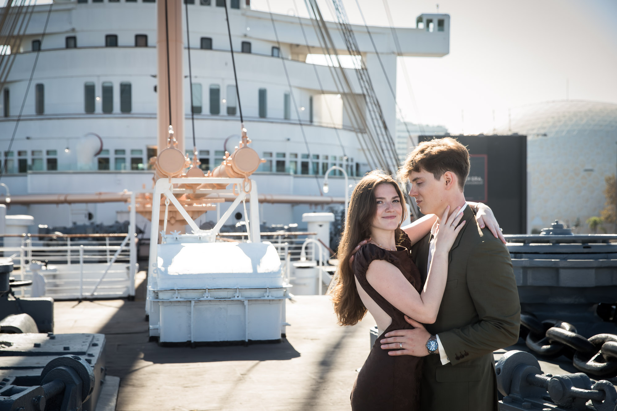 Couple embracing on the ship's deck as she looks at the camera, with the white superstructure and dome behind them, Queen Mary photo shoot