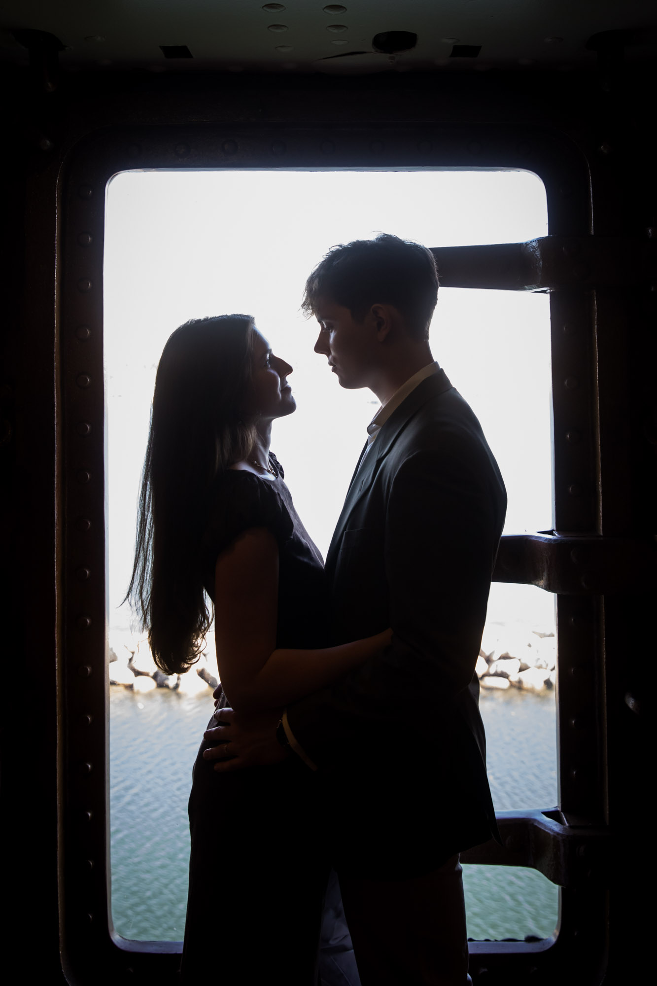 Silhouette of a couple facing each other in a ship's riveted doorway with the harbor behind them, by a Los Angeles photographer