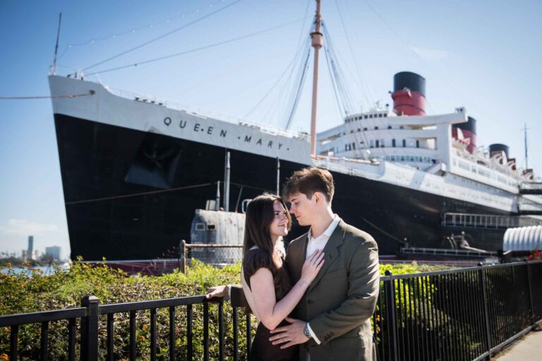 Engaged couple embracing along the waterfront fence with the Queen Mary ocean liner behind them, Queen Mary photo shoot
