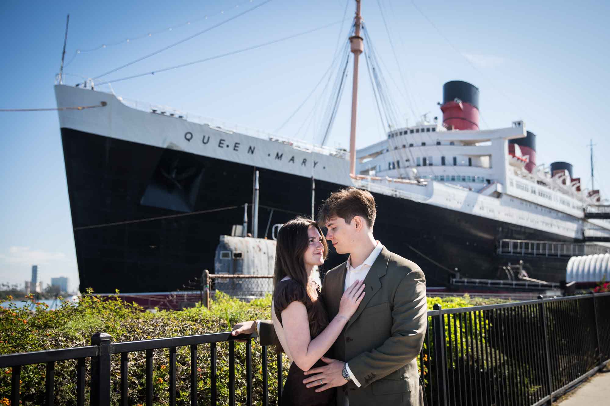 Engaged couple embracing along the waterfront fence with the Queen Mary ocean liner behind them, Queen Mary photo shoot