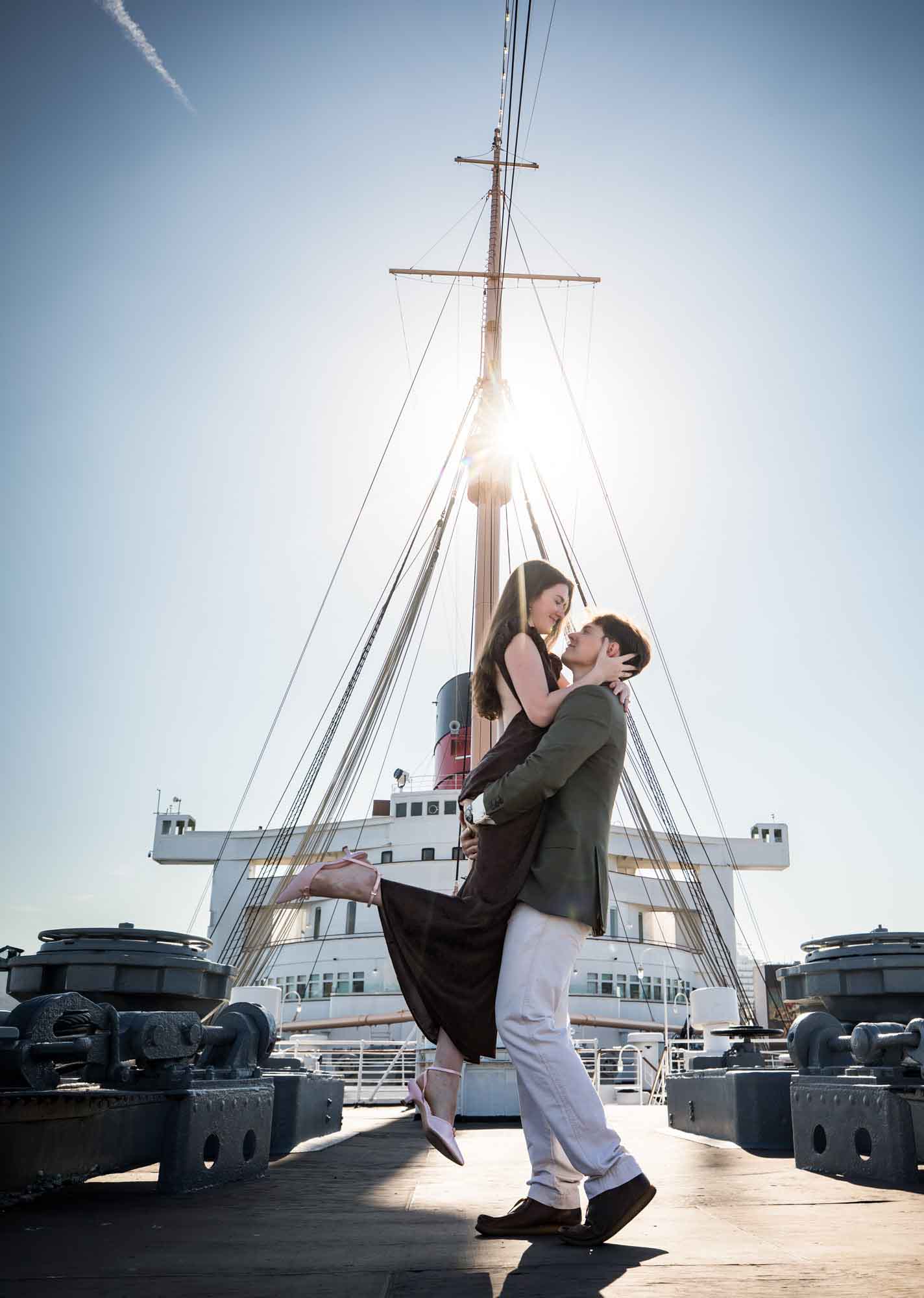 Man lifting his fiancée in the air as the sun flares behind the ship's mast, Queen Mary photo shoot