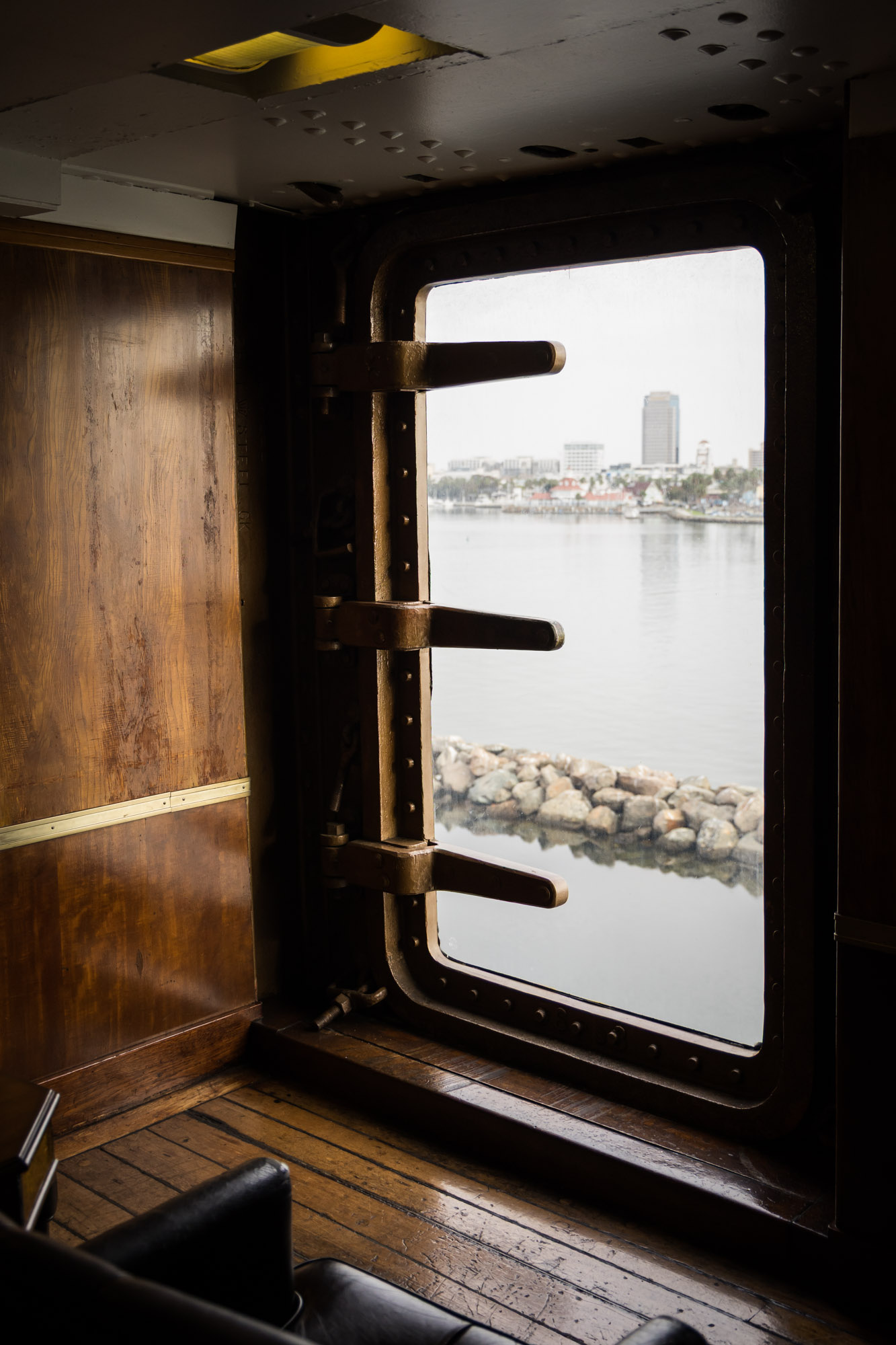Riveted porthole door with latches framing the harbor view beside warm wood paneling, a Queen Mary photo shoot interior detail