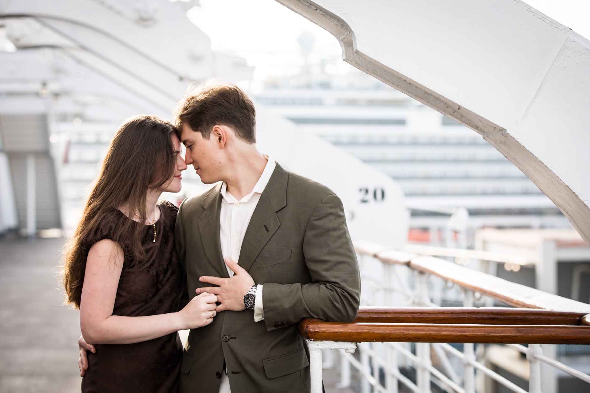 Couple touching foreheads at the rail with a cruise ship behind them, a romantic moment from a Queen Mary photo shoot
