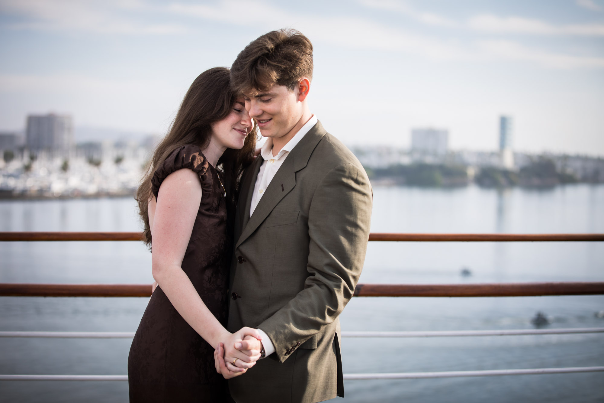 Woman resting her head against the man's as they hold hands at the rail during a Queen Mary photo shoot, with the Long Beach harbor behind them
