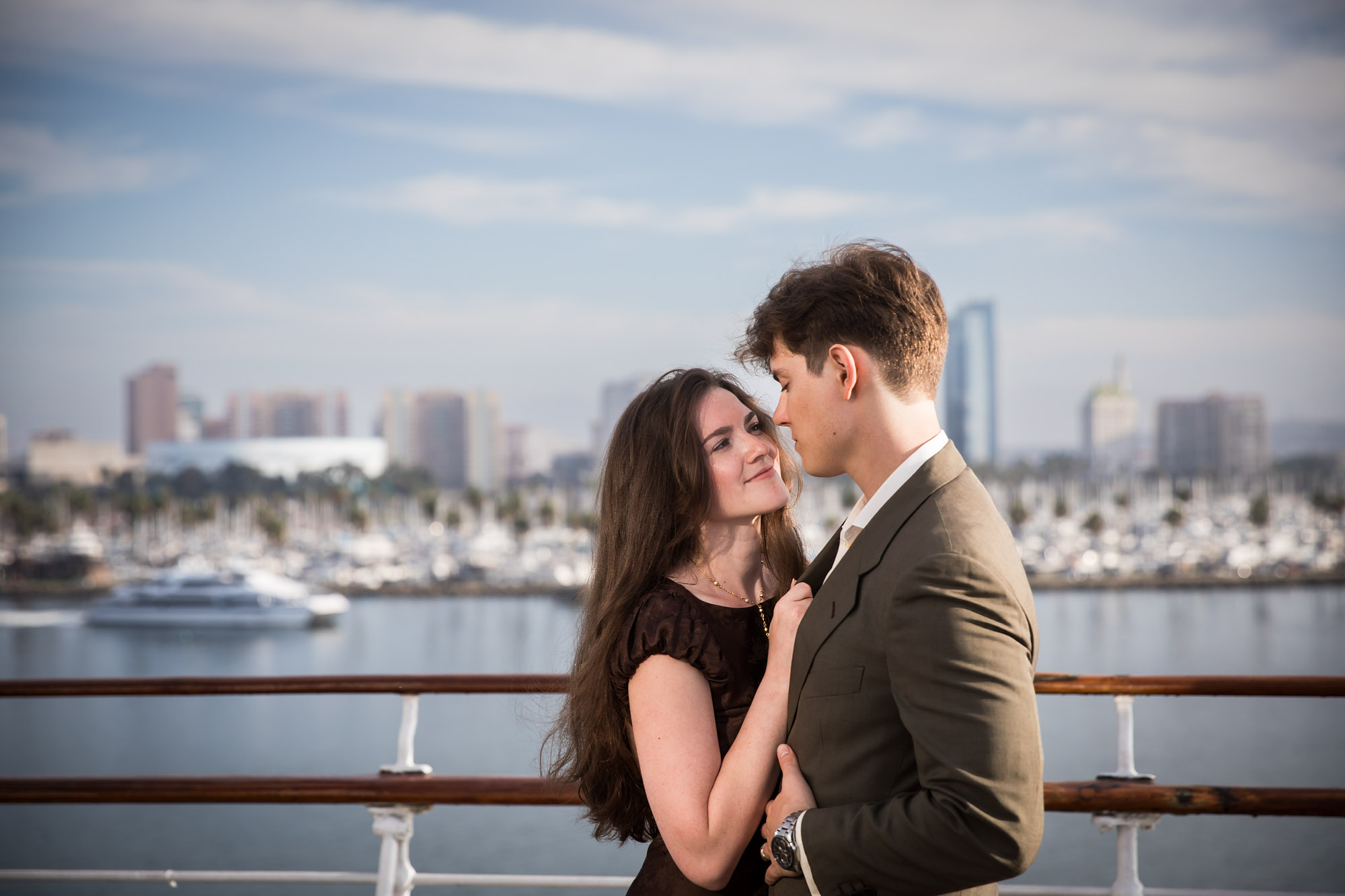 Woman looking up at the man with her hand on his lapel and a yacht in the harbor behind, during their Queen Mary photo shoot