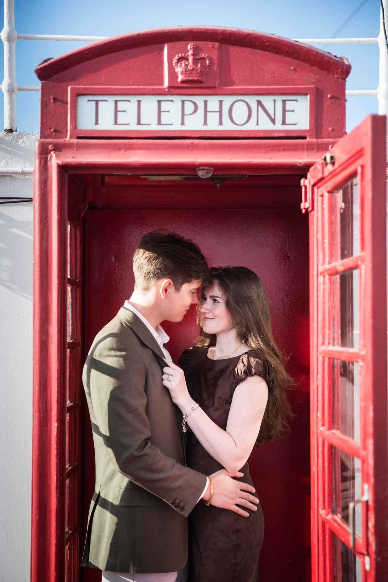 Couple touching foreheads inside a red British-style telephone box, a charming stop on a Queen Mary photo shoot